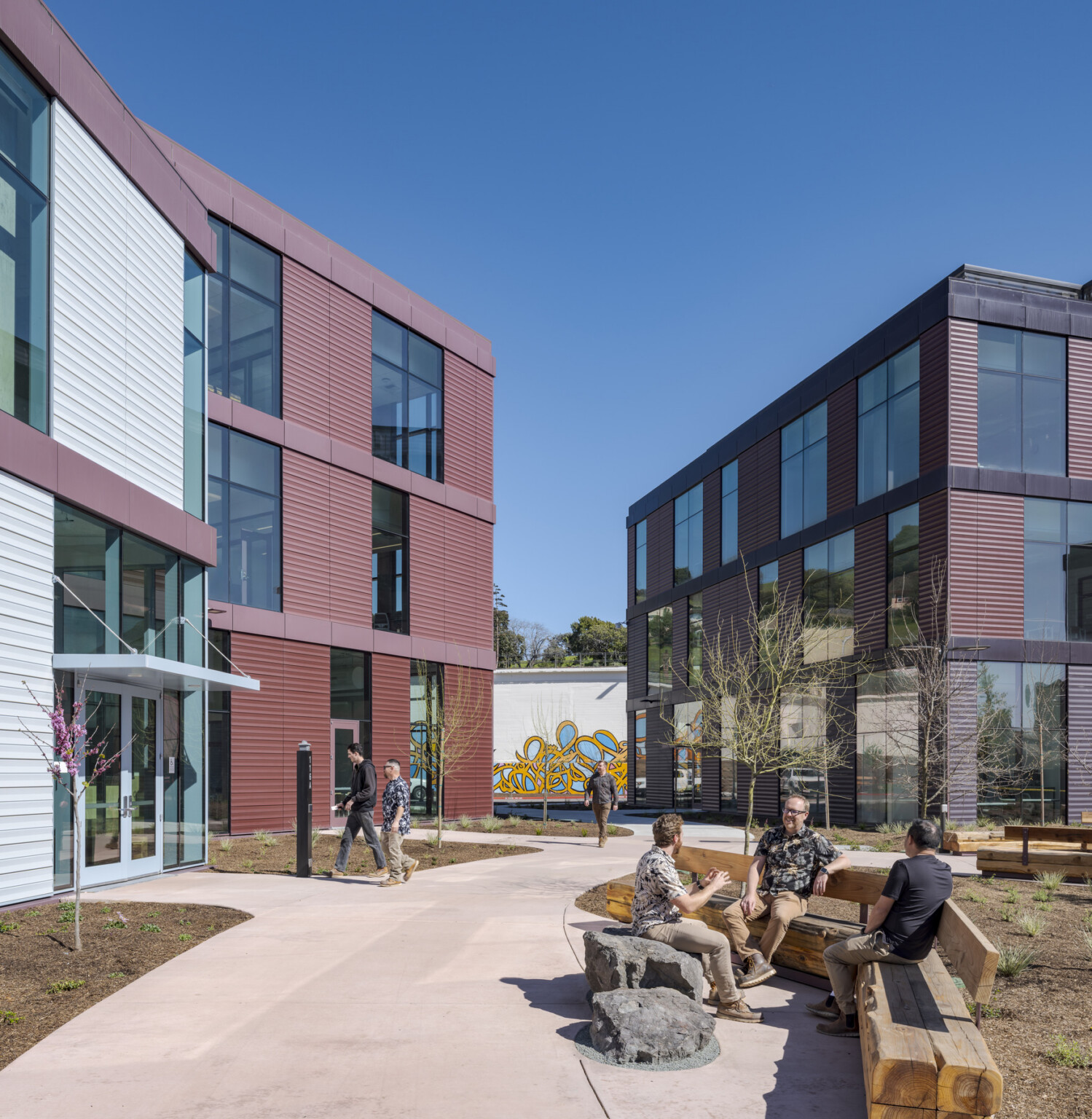 A clear blue sky above an outdoor courtyard situated between two maroon-colored buildings; several people are seated on wooden benches near one of the building entrances, enjoying the open-air setting.