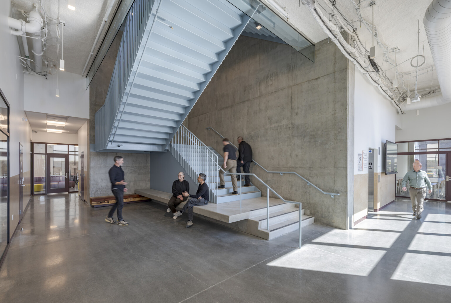 A spacious hallway with a high ceiling and a prominent staircase; several people walk through the space or sit on a built-in cement bench attached to the staircase.