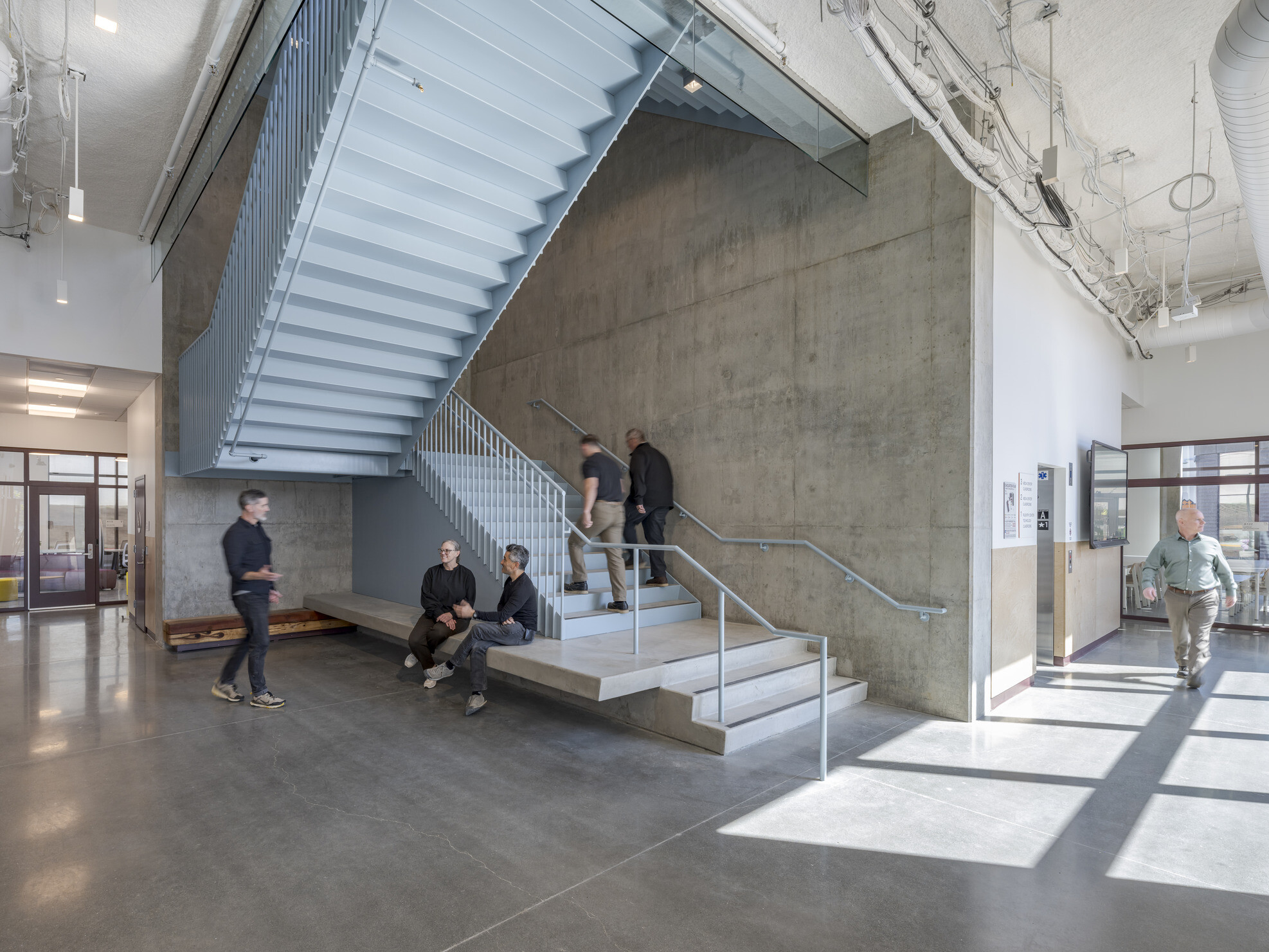 A spacious hallway with a high ceiling and a prominent staircase; several people walk through the space or sit on a built-in cement bench attached to the staircase.