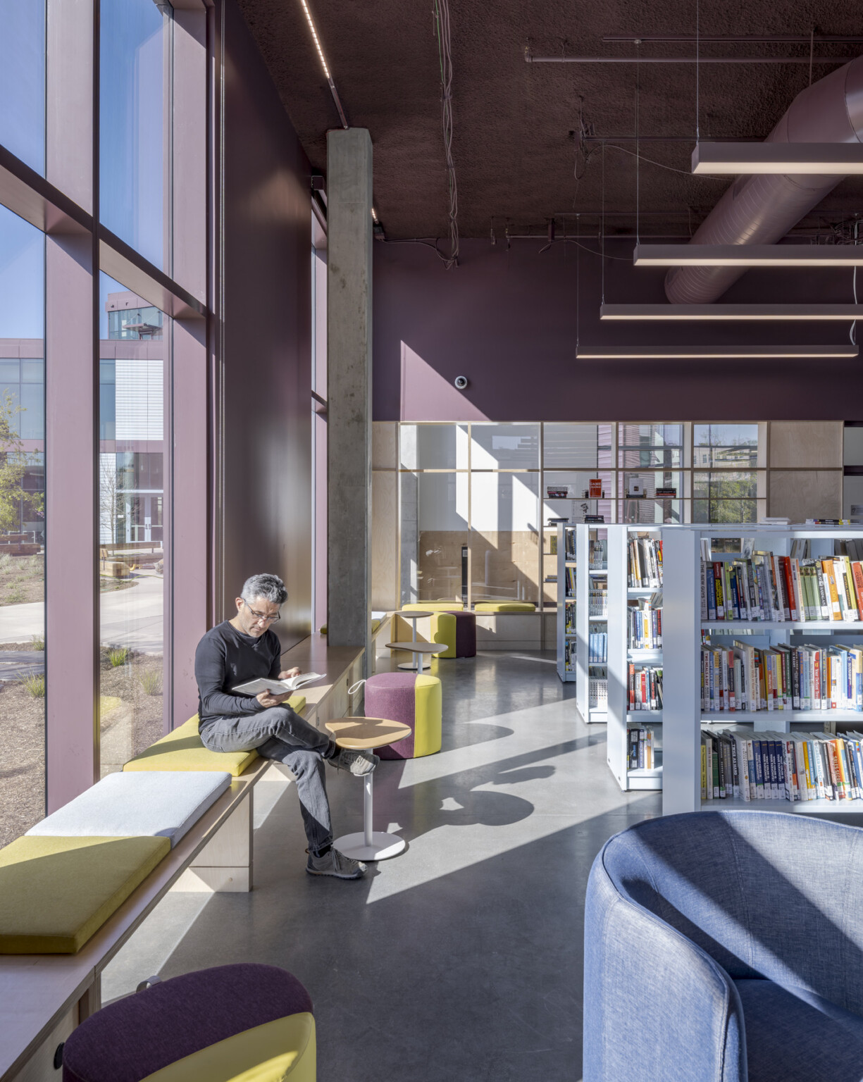 A man reads a book on a window bench bathed in sunlight beside floor-to-ceiling windows, surrounded by bookshelves in the center’s new library.