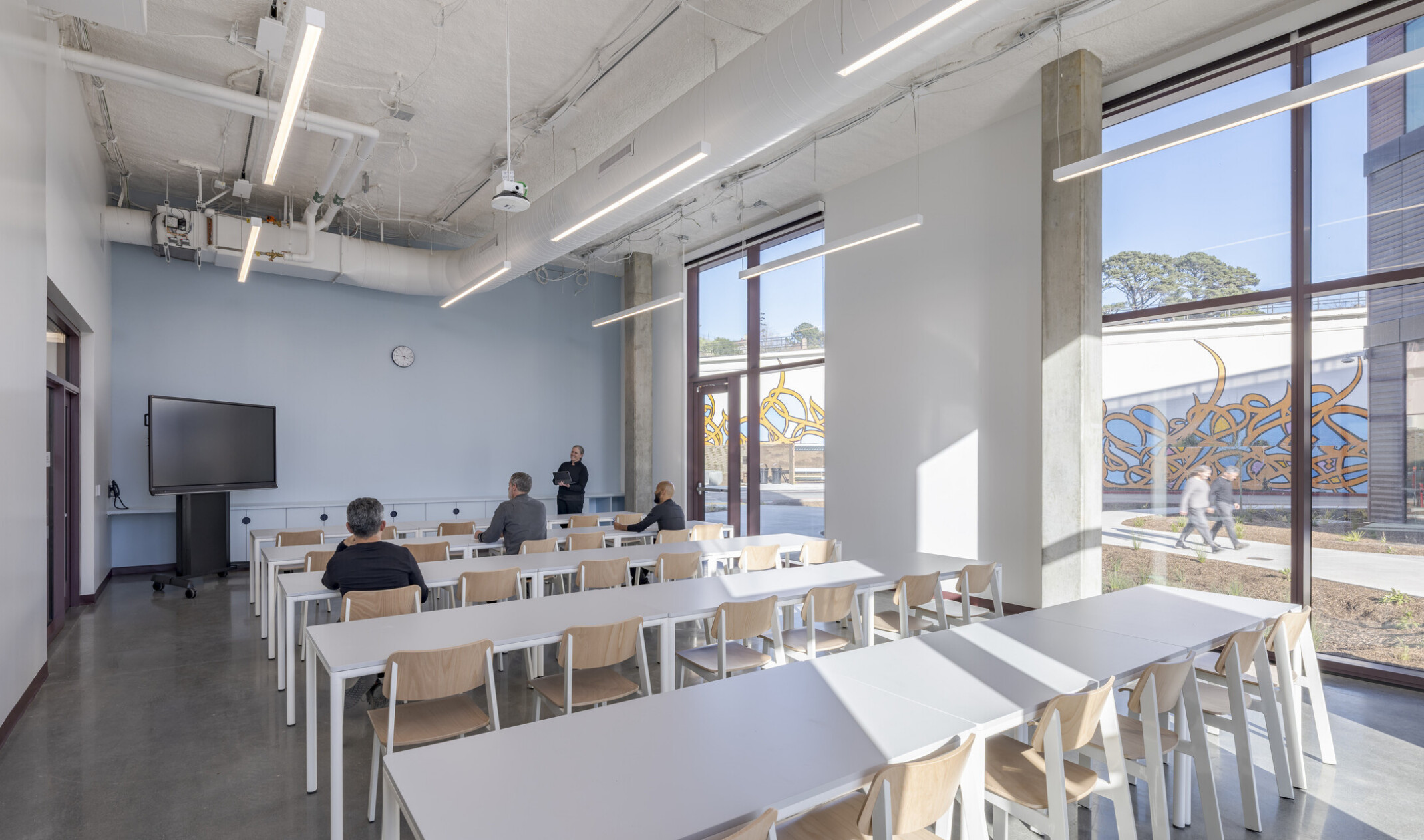 Bright classroom with long desk tables and ample seating; floor-to-ceiling windows reveal outdoor foot traffic and custom graffiti artwork on exterior concrete walls. An instructor stands at the front presenting to seated attendees.