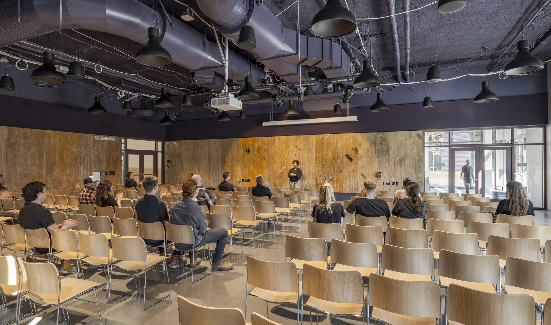 Theater-style room with rows of chairs filled by a medium-sized audience, while a speaker addresses the group from the front, standing before wooden paneling.