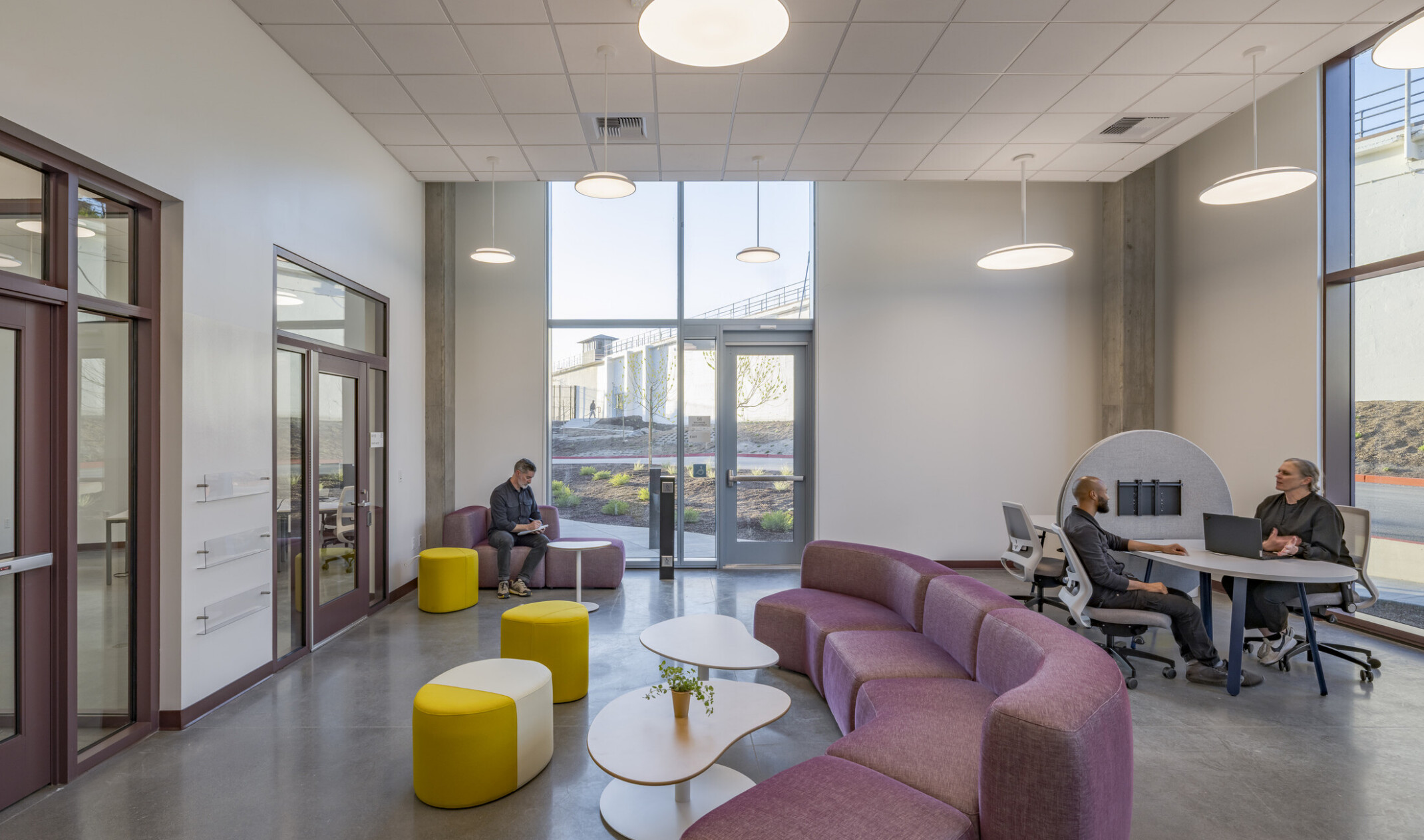Multipurpose room furnished with a long maroon couch, modern coffee tables, colorful stools, and a desk area; windows let in natural sunlight and offer direct views into the natural landscape and courtyard outside.