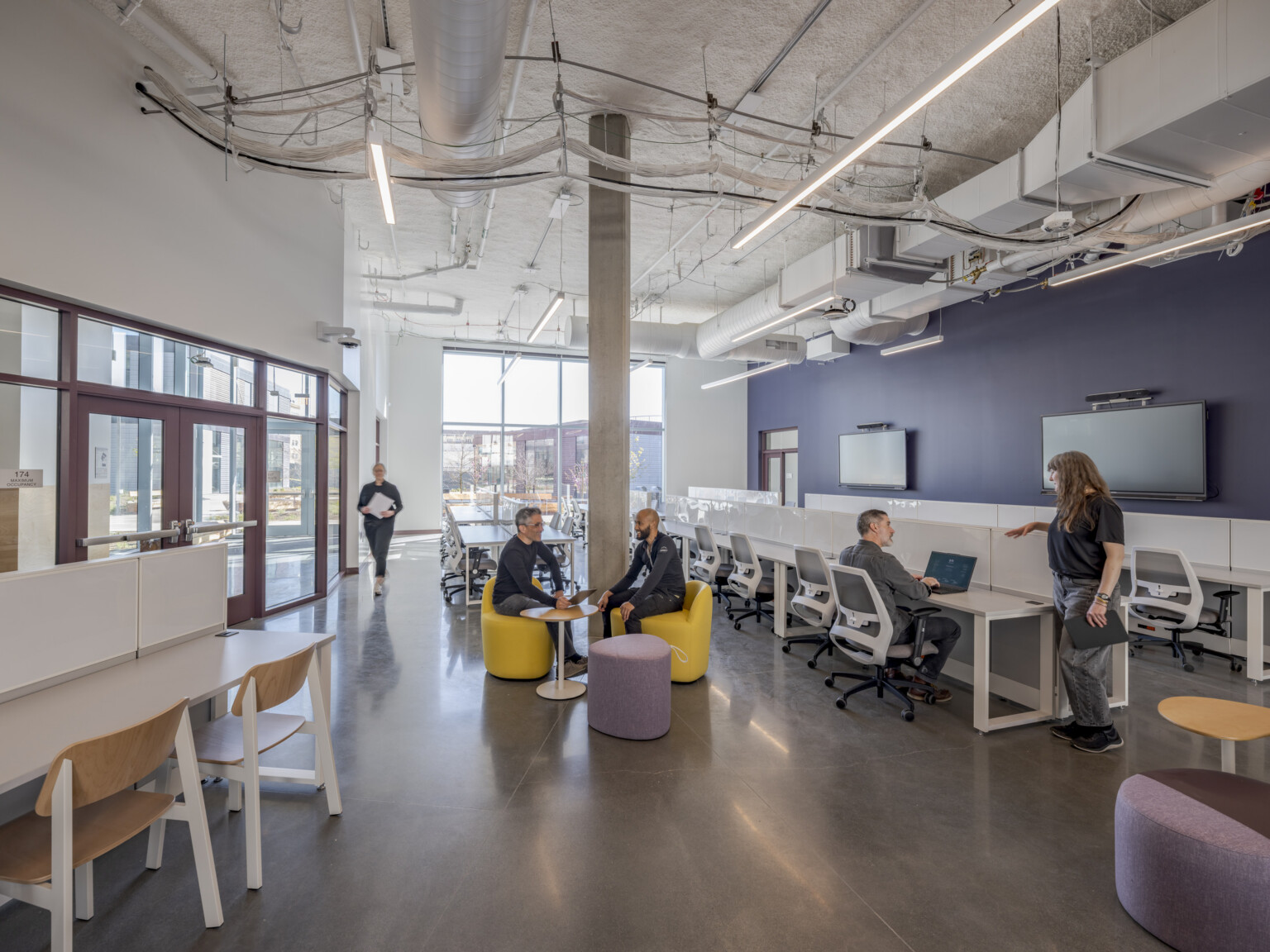 A learning lab with high ceilings and multiple work areas, where people sit on stools and at desks; floor-to-ceiling windows in the background provide direct views into the center’s courtyard.