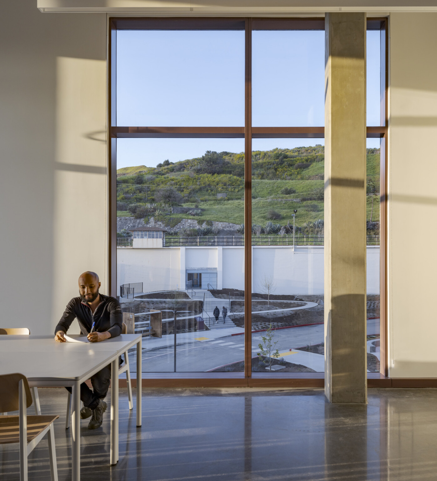 A man sits at a table inside, with floor-to-ceiling windows behind him offering a view of the green hills encircling the facility.