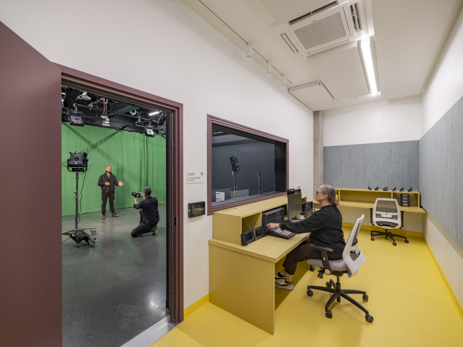 A performer is filmed in a studio in front of a green screen by a cameraman; through an open doorway, a sound engineer is visible at a sound booth, observing from an adjacent room.