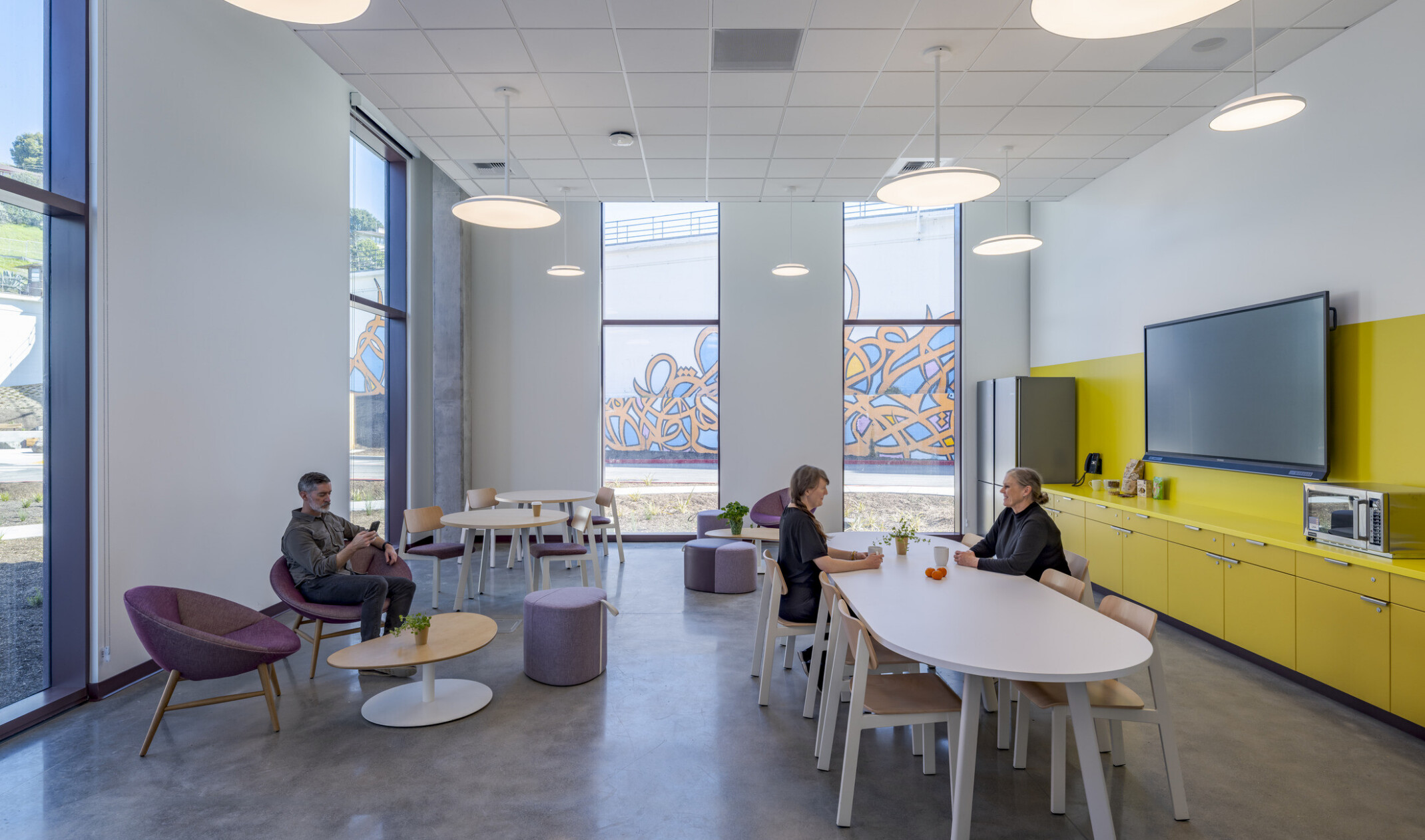 Sitting room with several tables, chairs, counters, a fridge, and a mounted television, occupied by a few people; floor-to-ceiling windows provide a view of the courtyard and walkways outside.