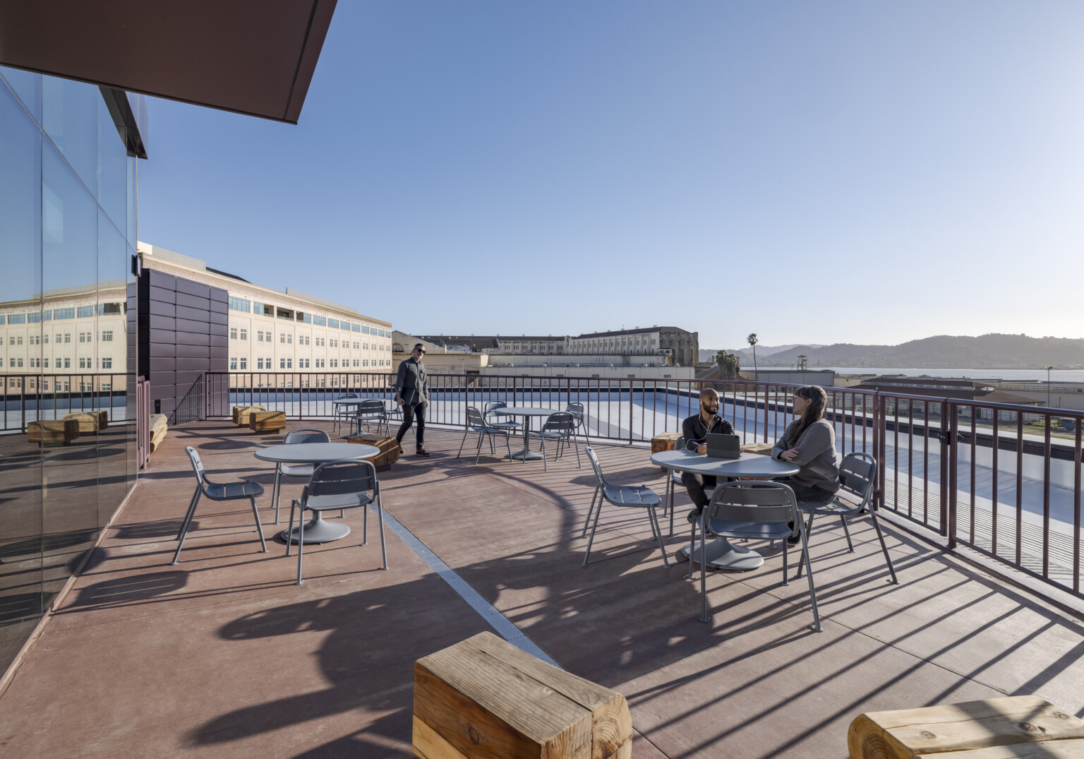 A balcony filled with tables and seating, where people sit and walk; the San Quentin facility appears in the immediate background, with Northern California hills rolling beyond.