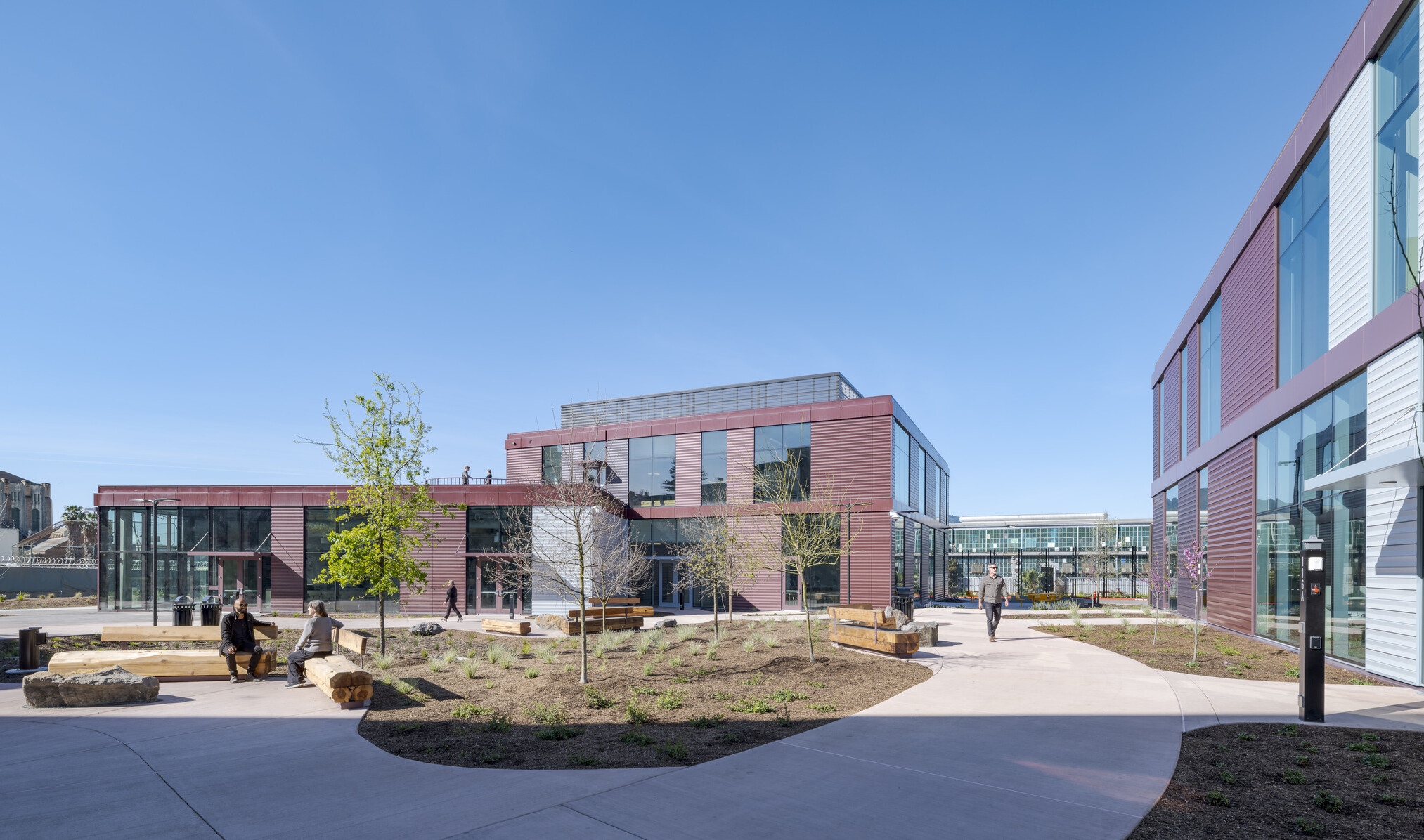 Wide shot showing two of the three new center buildings in the background, with a courtyard in the foreground featuring wide walkways, wooden benches, natural landscaping, and people sitting or walking throughout.