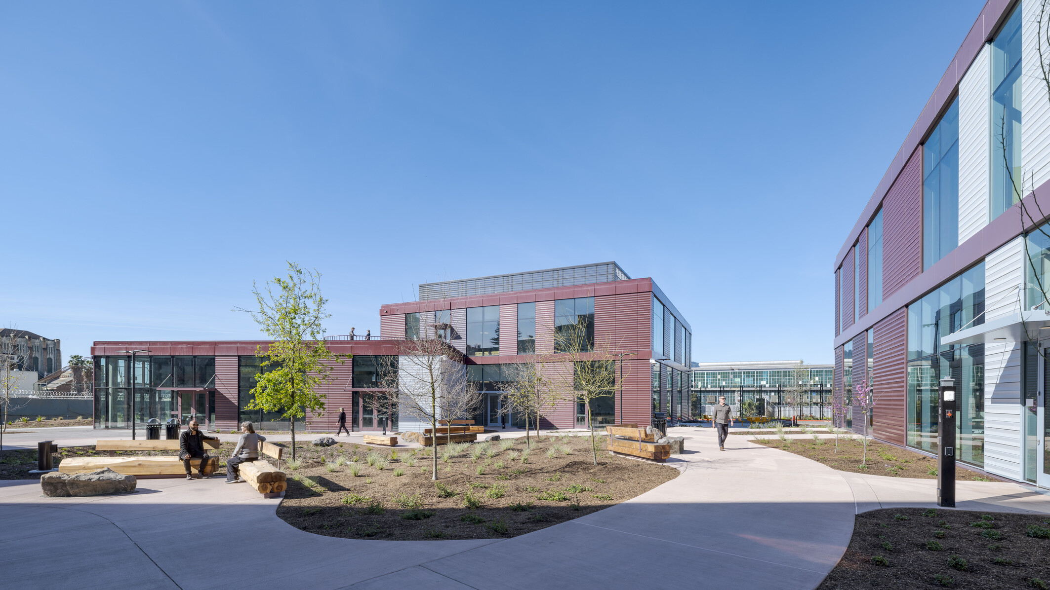 Wide shot showing two of the three new center buildings in the background, with a courtyard in the foreground featuring wide walkways, wooden benches, natural landscaping, and people sitting or walking throughout.