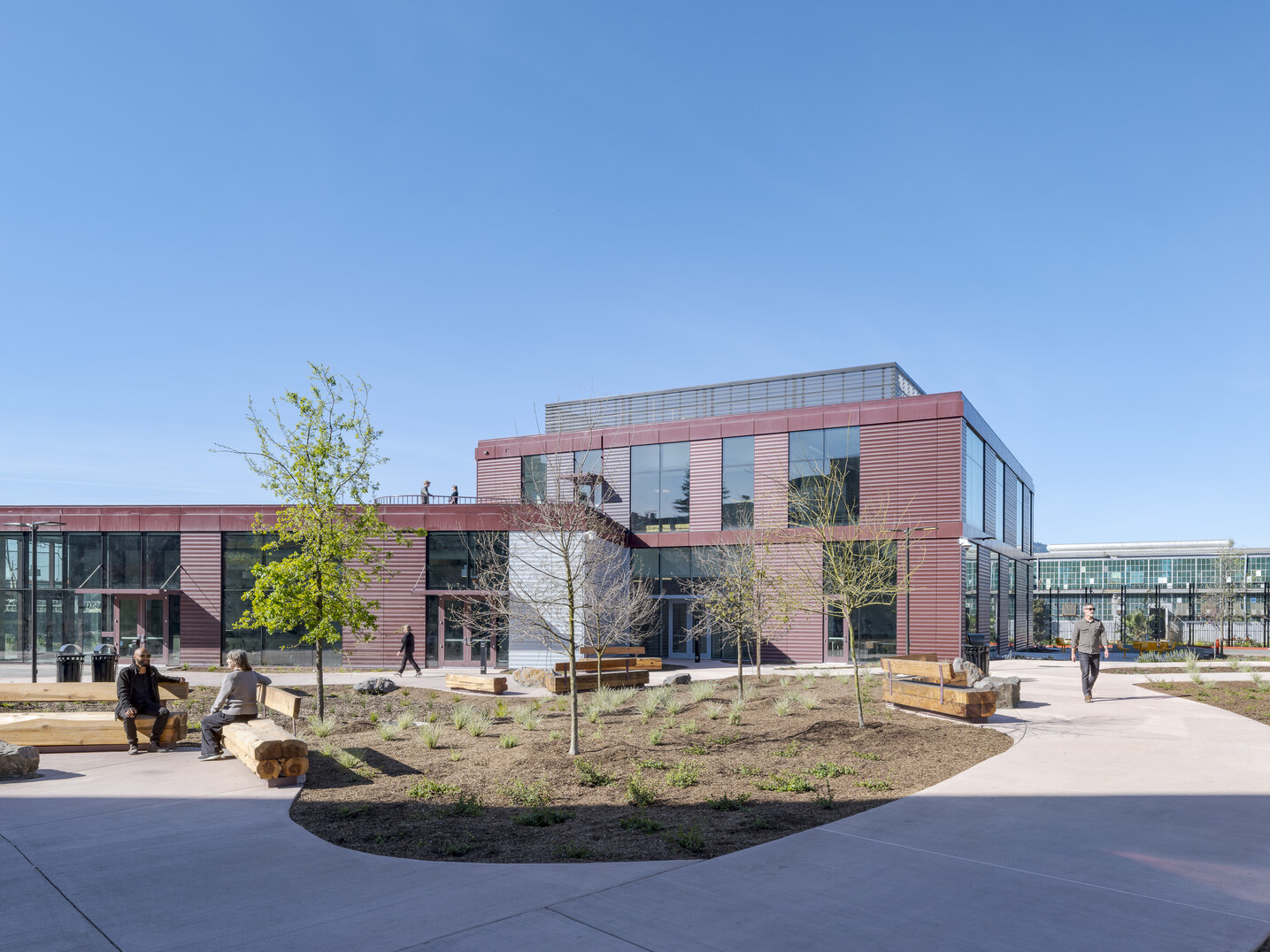 Wide shot showing two of the three new center buildings in the background, with a courtyard in the foreground featuring wide walkways, wooden benches, natural landscaping, and people sitting or walking throughout.
