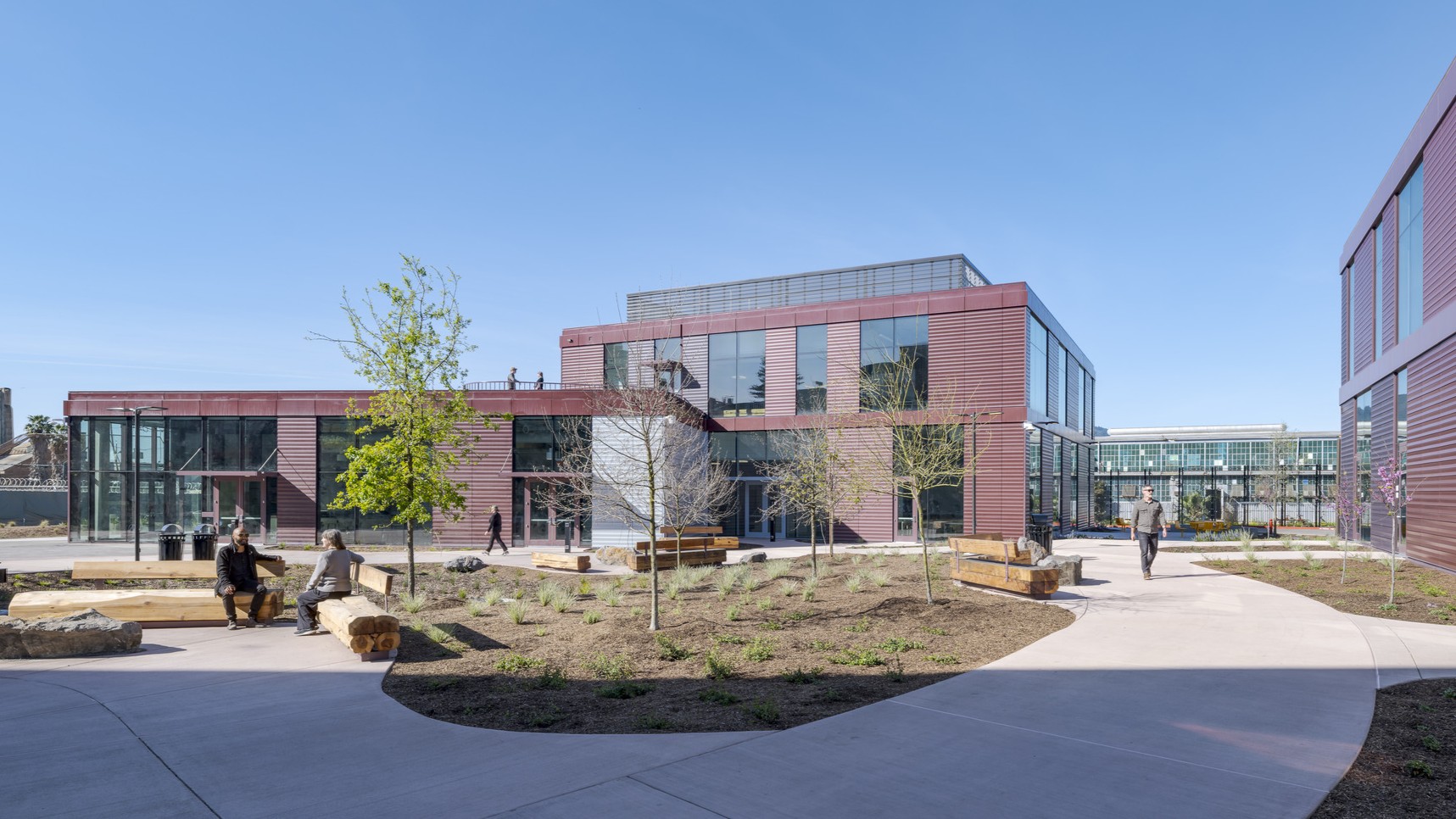 Wide shot showing two of the three new center buildings in the background, with a courtyard in the foreground featuring wide walkways, wooden benches, natural landscaping, and people sitting or walking throughout.