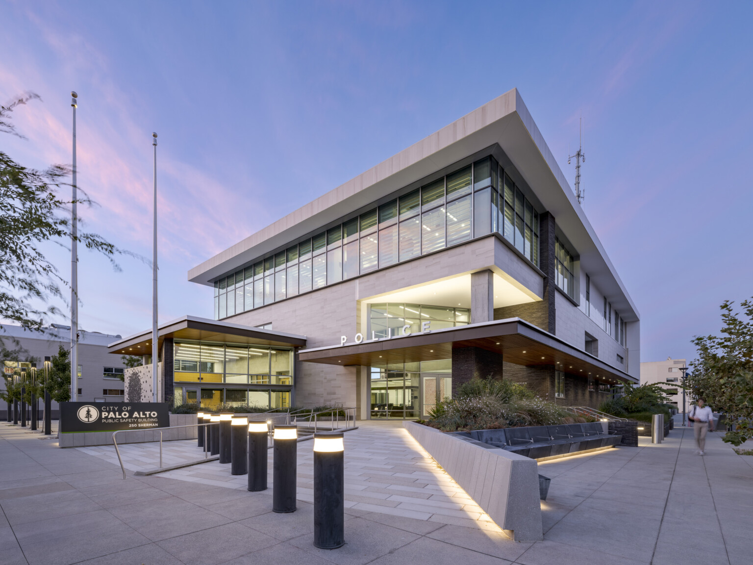 Evening shot of Palo Alto Public Safety Building & Emergency Operations Center; 2 story building fronted by a public plaza and covered entrance with a row of floor-to-ceiling windows illuminated along the top floor