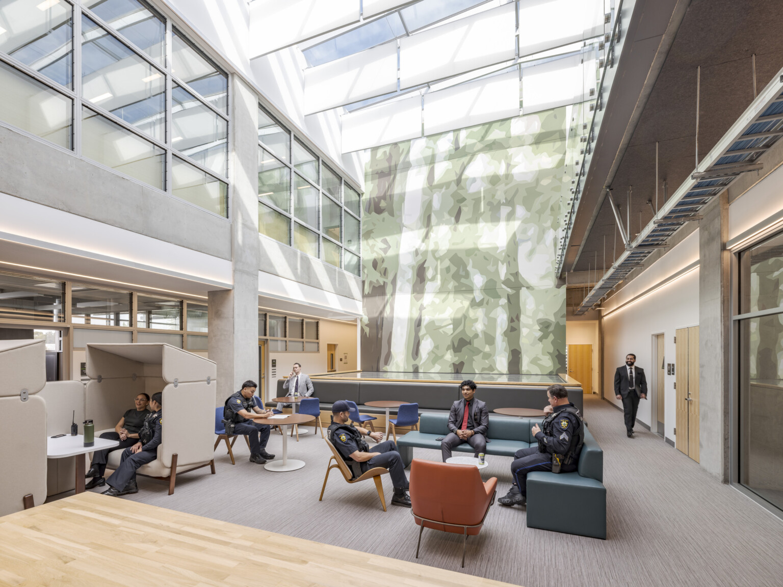 Palo Alto Public Safety Building & Emergency Operations Center; Detectives in suits and uniformed police officers sit in conversation at café tables and soft seating in a double-height atrium that is bathed in sunlight. An abstract mural of a tree covers the back wall