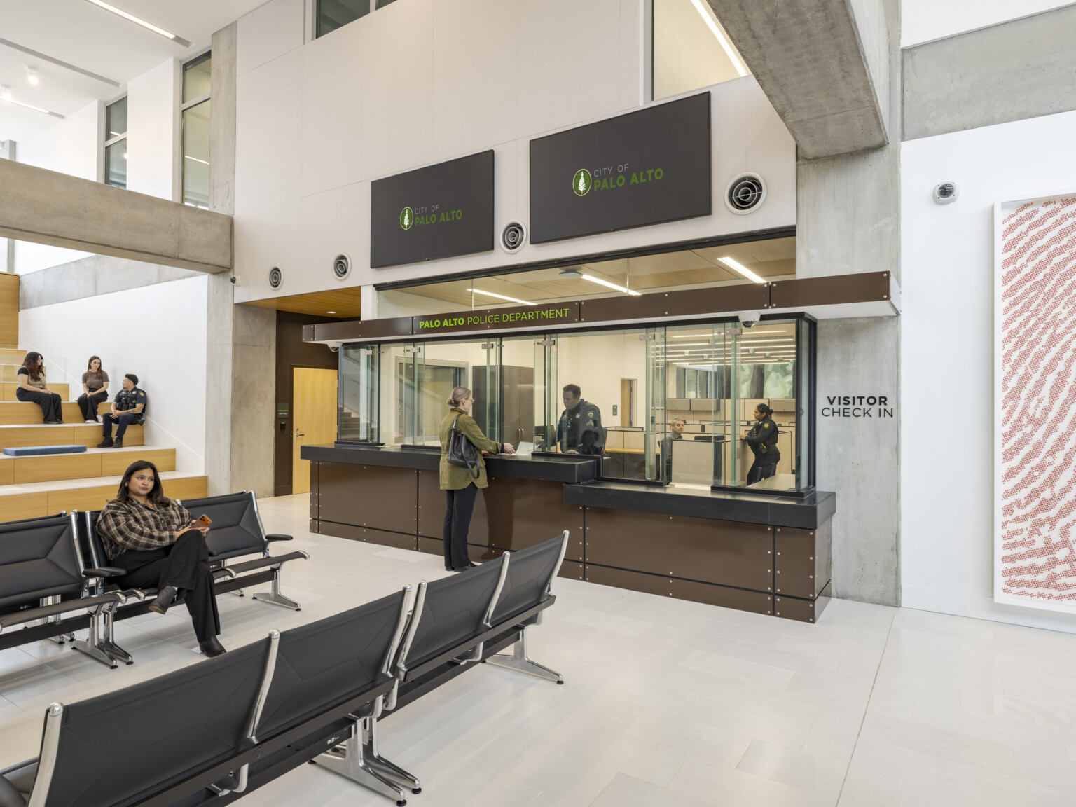 Palo Alto Public Safety Building & Emergency Operations Center; a woman at the visitor check-in counter speaks to a uniformed officer behind the glass. Behind the officer is a workspace with low cubicles. In the public waiting area, a woman sits in a row of seats, while behind her, two visitors and a police officer chat on wooden amphitheater-style benches