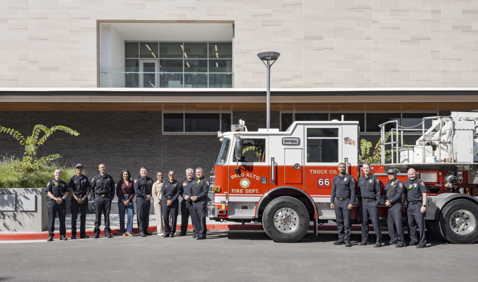 group of 13 smiling fire department personnel stand flanking the cab of PAFD Engine 66, which is parked in front of Palo Alto Public Safety Building & Emergency Operations Center
