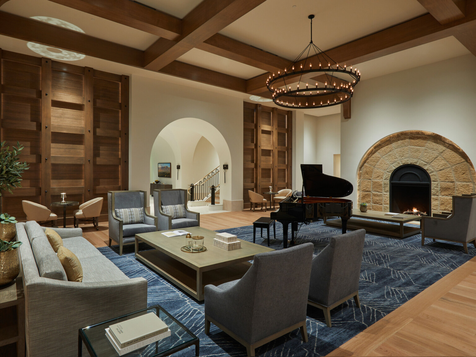 Elegant seating area with a grand piano, blue rug, arched stone fireplace, and wooden ceiling beams