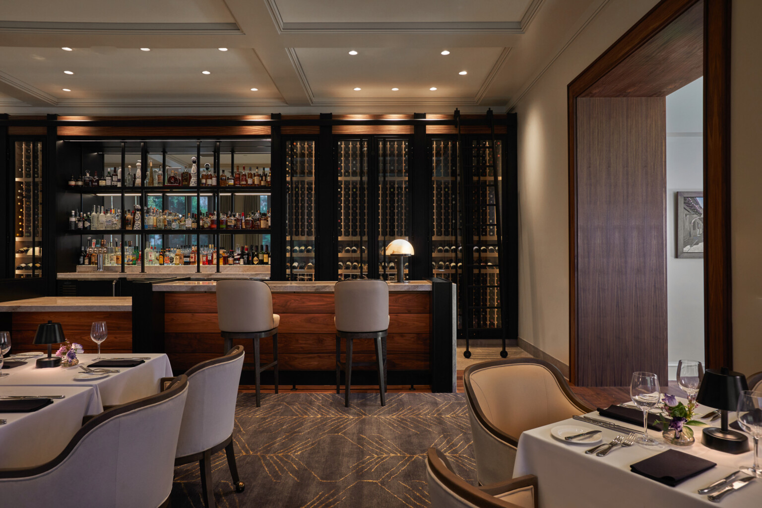 Elegant restaurant interior with a bar, wine racks, and dining tables set with white tablecloths and floral centerpieces.
