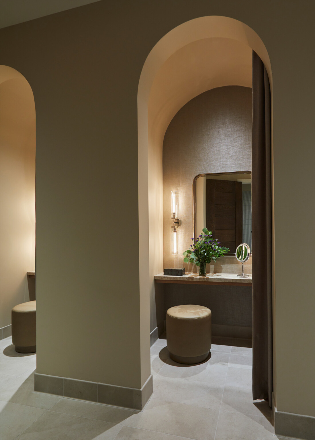 Alcove with a counter with a vanity mirror and plants on top with a light fixture and mirror above, and stools in front in warm beige tones