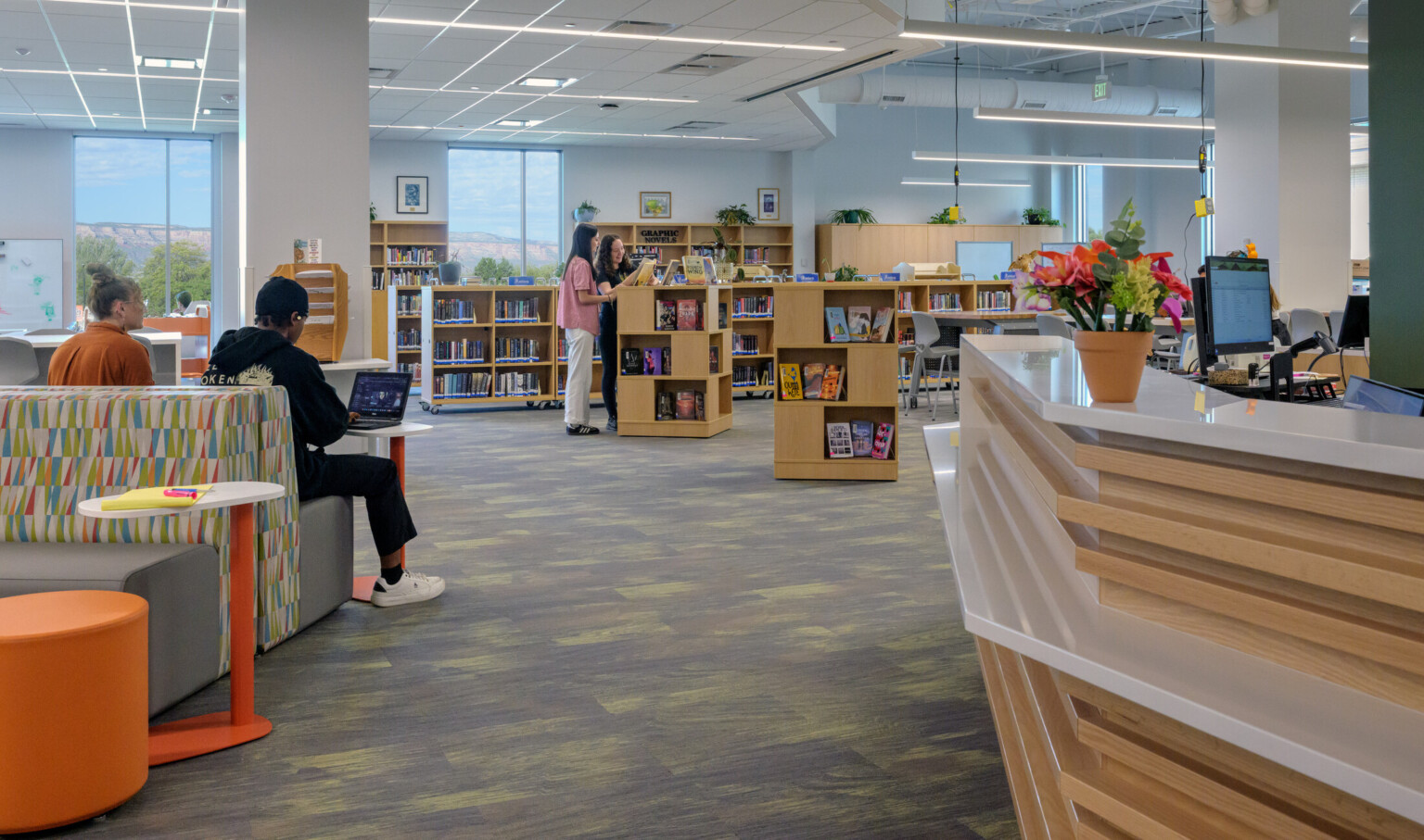 modern and bright library with students working and reading in comfy benches; students at book stand look at displayed books; librarian desk counter to the right