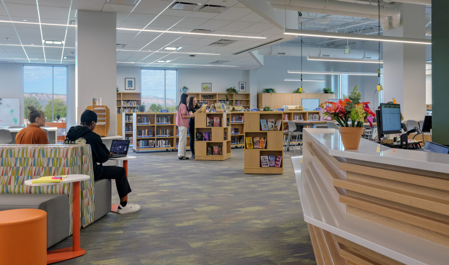modern and bright library with students working and reading in comfy benches; students at book stand look at displayed books; librarian desk counter to the right