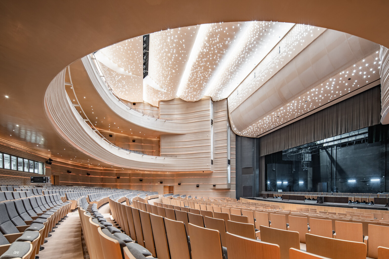 Modern auditorium with semi-circular beige seating, wave-like wooden ceiling, starry lighting, and a large stage with curtain.