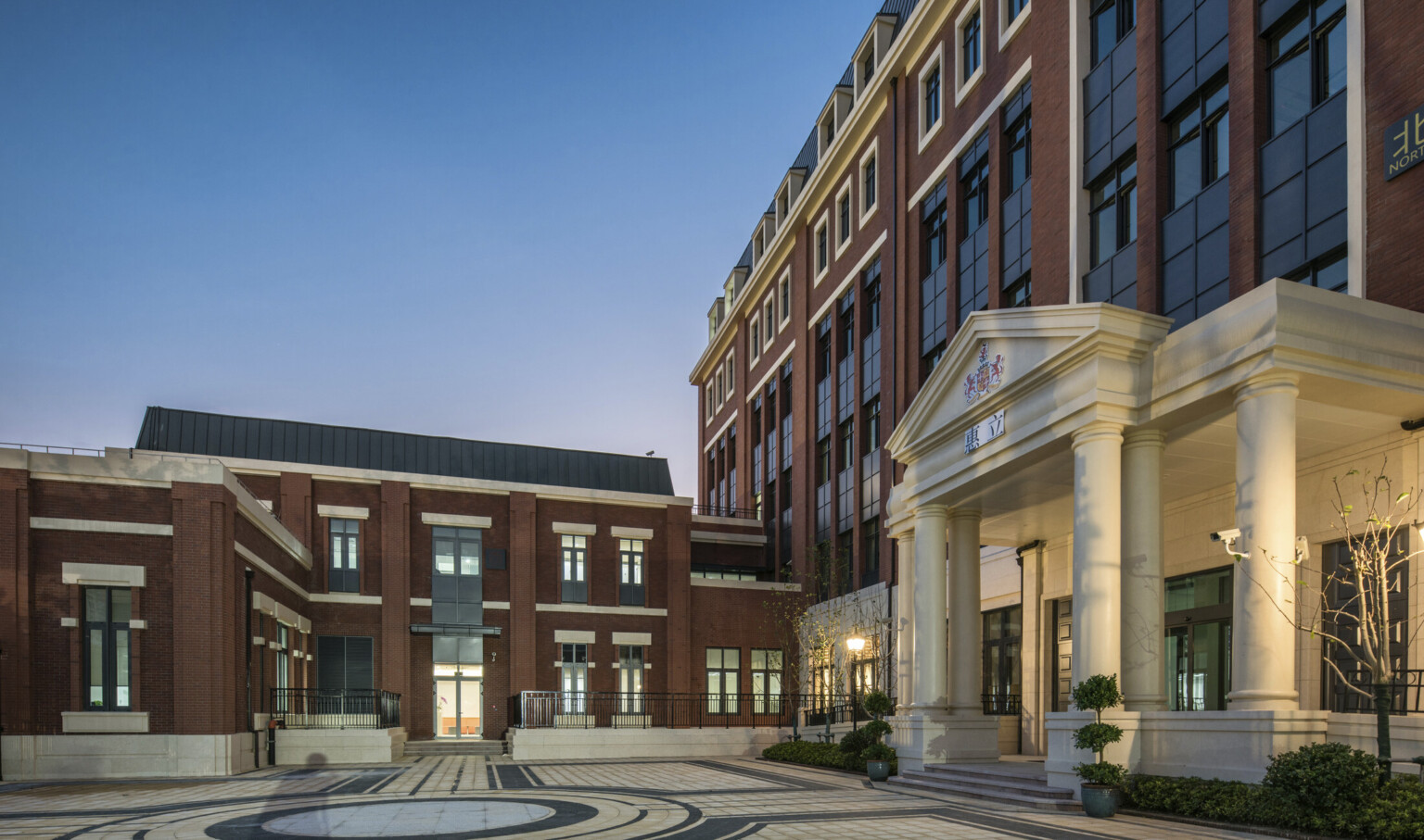Courtyard view of white stone front entry with framed with columns around glass doors. 2 story brick building connects left
