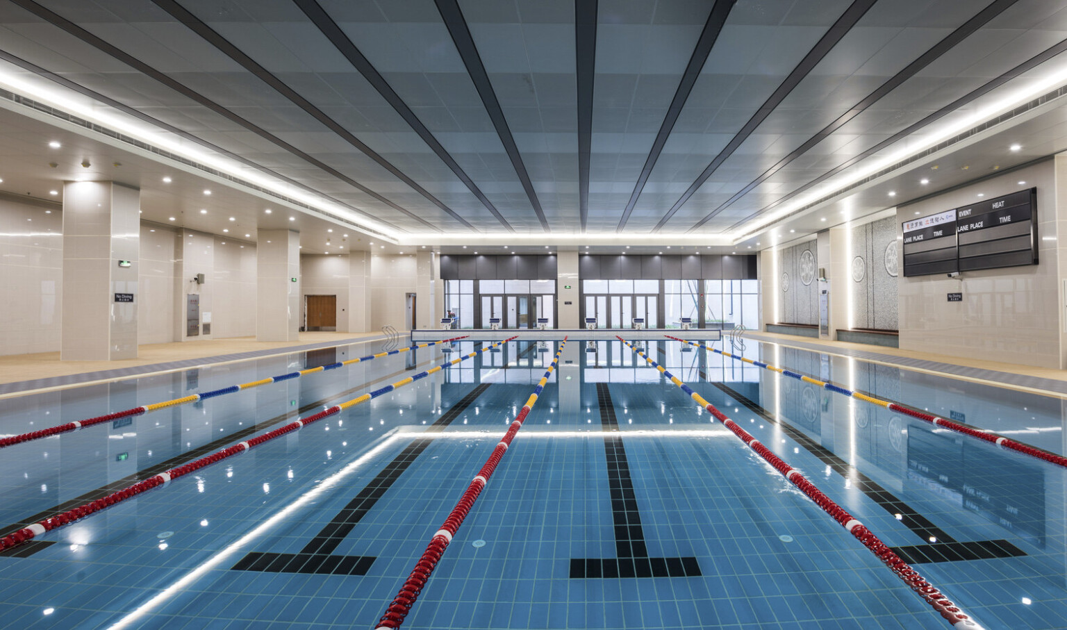 View looking down a lap pool to floor to ceiling windows. Recessed lighting lines the pool from above and tiled path on sides