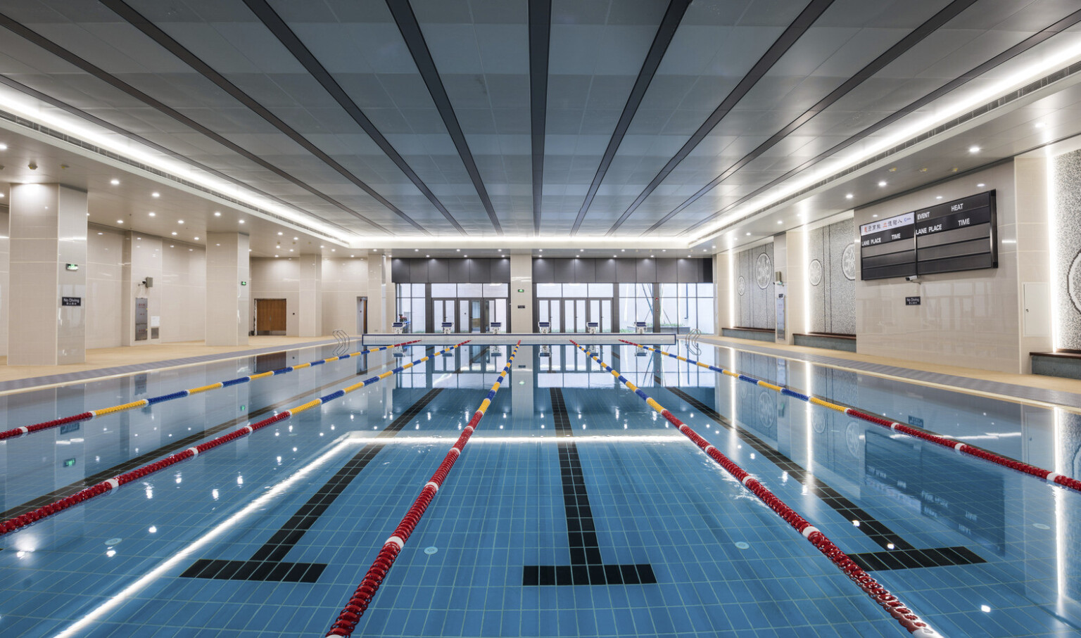 View looking down a lap pool to floor to ceiling windows. Recessed lighting lines the pool from above and tiled path on sides