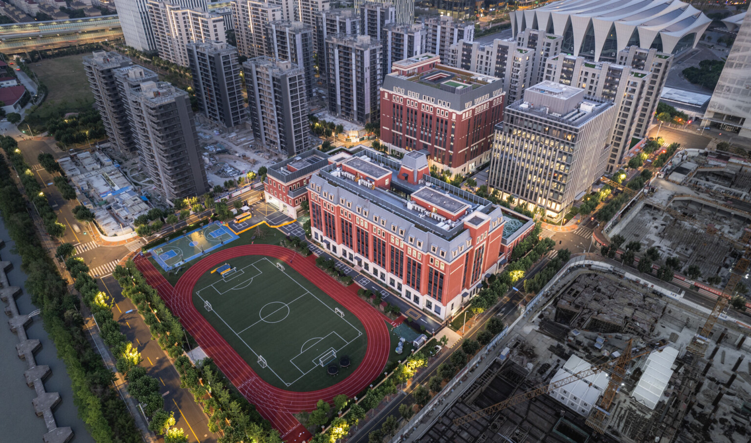 Aerial view of a red-brick building complex with a soccer field surrounded by a running track and adjacent high-rise buildings.