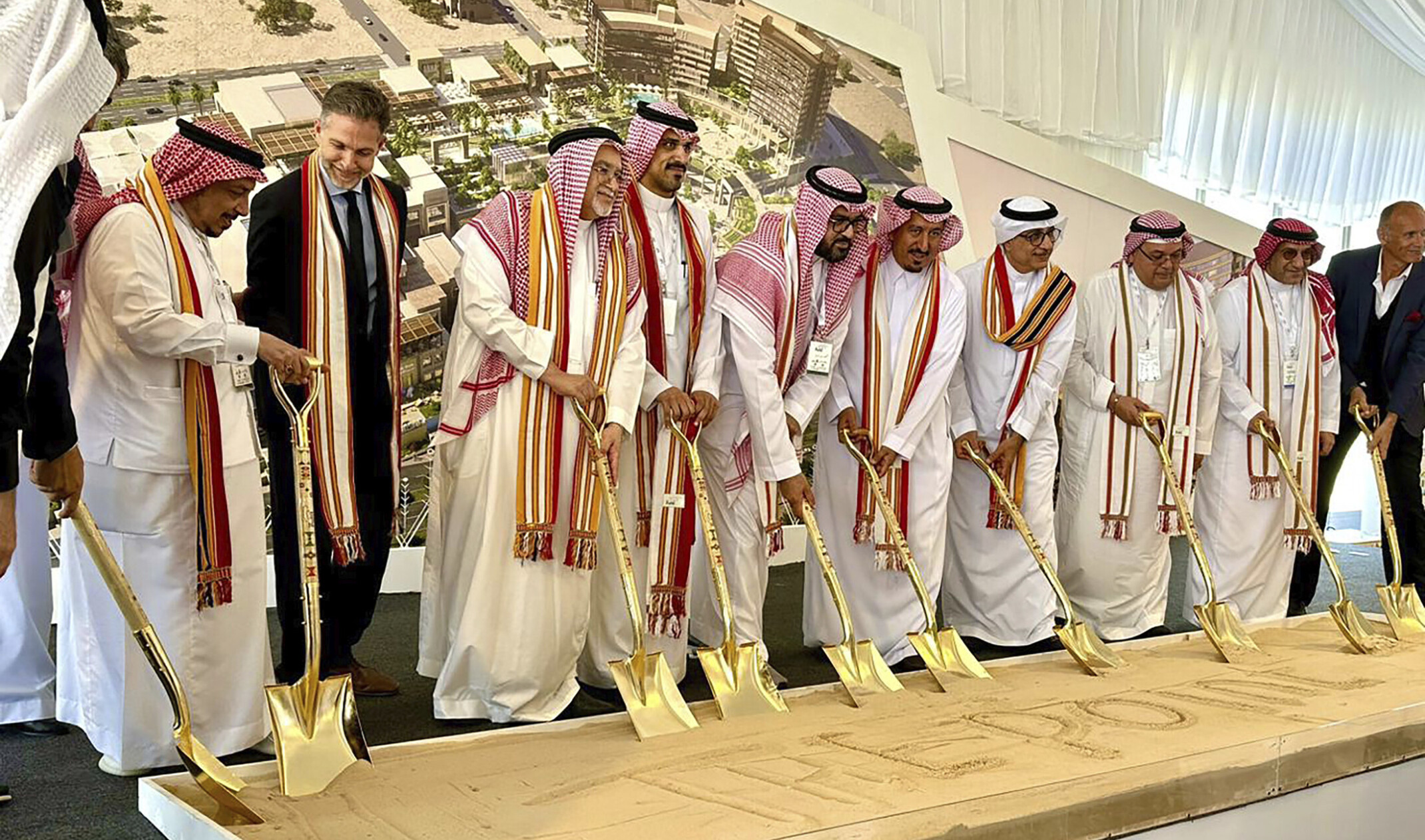 Multiple men wearing white robes at a ground breaking holding gold shovels digging into a box of sand