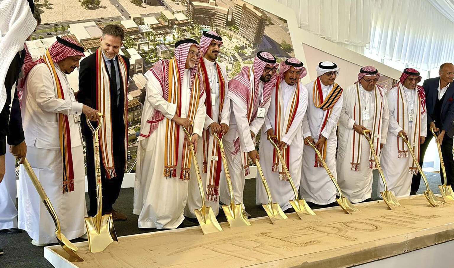 Multiple men wearing white robes and colorful scarves at a ground breaking holding gold shovels digging into a box of sand