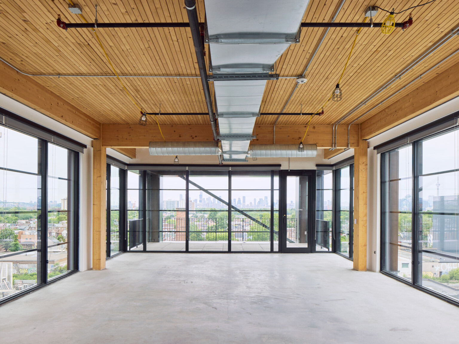 Large room with exposed wooden beams, floor to ceiling black framed windows overlooking a skyline filled with large buildings