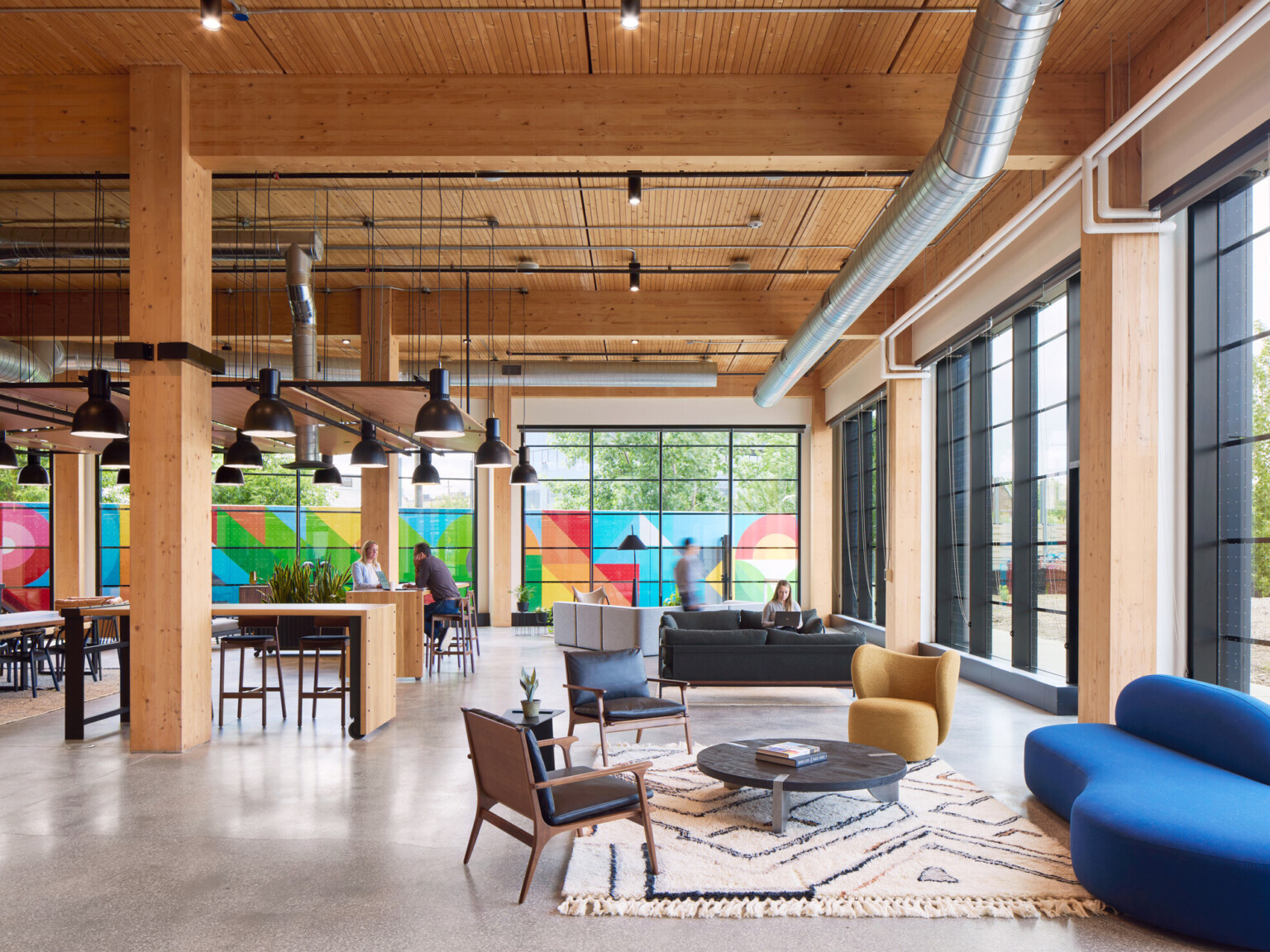 Large office space with exposed wooden beams, floor to ceiling black framed windows, and colorful mural outside