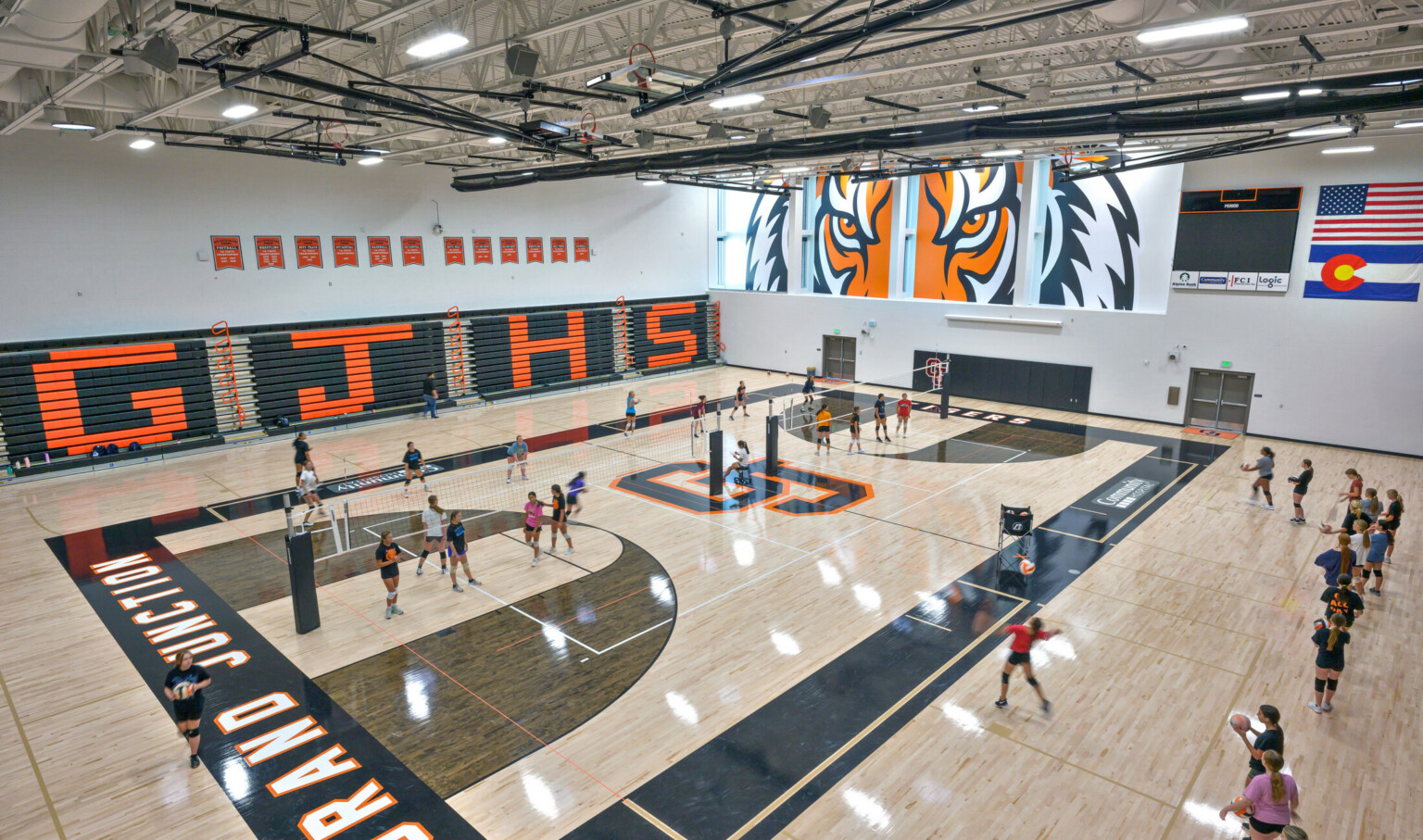 large elevated shot of high school volleyball practice; Grand Junction logo, orange and navy blue colors decorate the court; close up animated image of a tiger