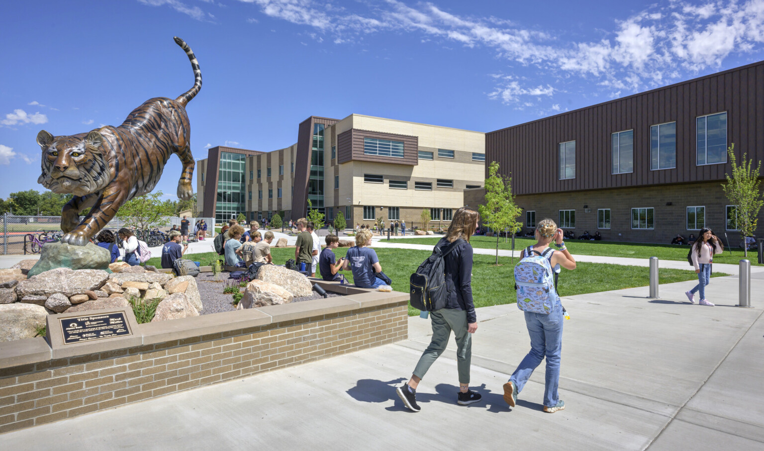 students walking into Grand Junction High School; Large brass tiger sculpture to the left sectioned out by bench perimeter; tiger is in a position of jumping down from a elevated height