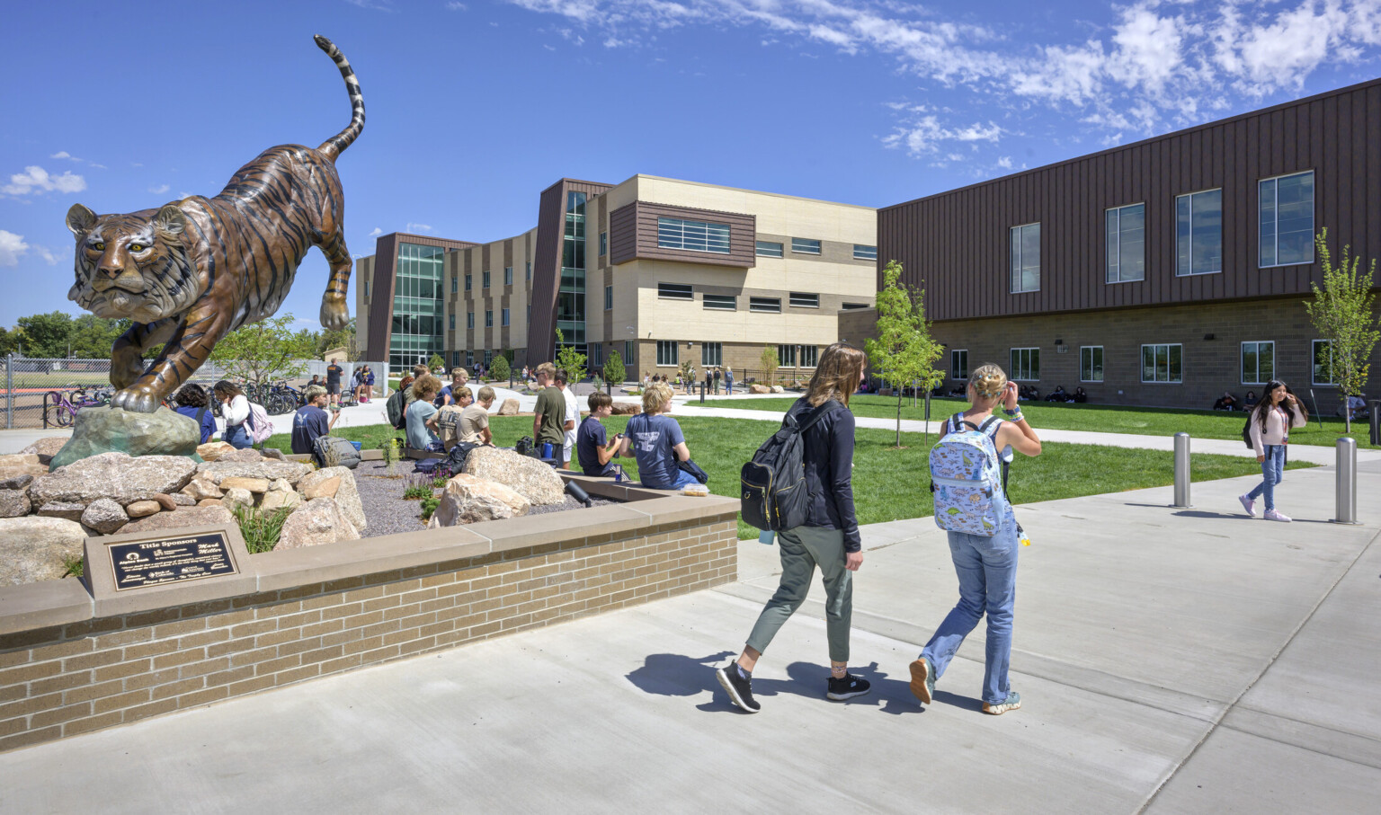 students walking into Grand Junction High School; Large brass tiger sculpture to the left sectioned out by bench perimeter; tiger is in a position of jumping down from a elevated height