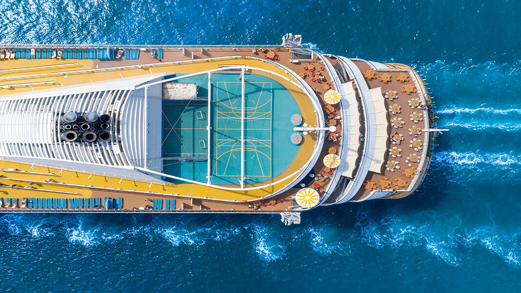 Aerial view of a cruise ship in blue water showing multiple decks with tables and chairs and a green playing court.