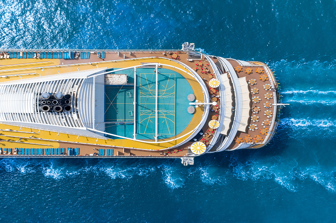 Aerial view of a cruise ship in blue water showing multiple decks with tables and chairs and a green playing court. Aerial view of a cruise ship in blue water showing multiple decks with tables and chairs and a green playing court.