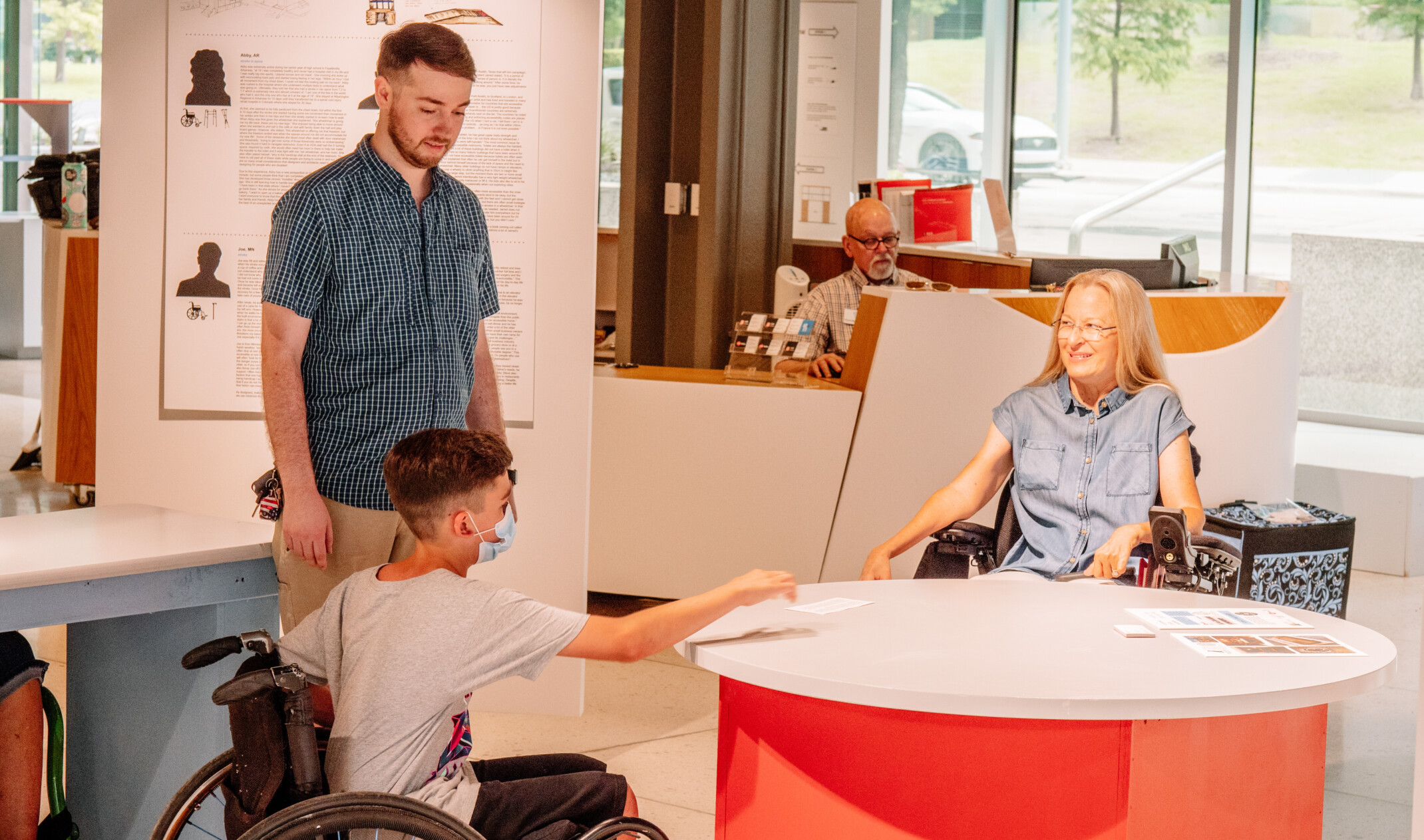 a child in a wheelchair leaning over an accessible height table with individuals. Red table with accessible signage.