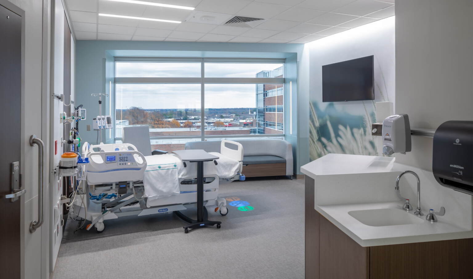 Hospital room with bed near large window. Mural on opposite wall with screen. Icons projected on floor at base of bed Hospital room with bed near large window. Mural on opposite wall with screen. Icons projected on floor at base of bed