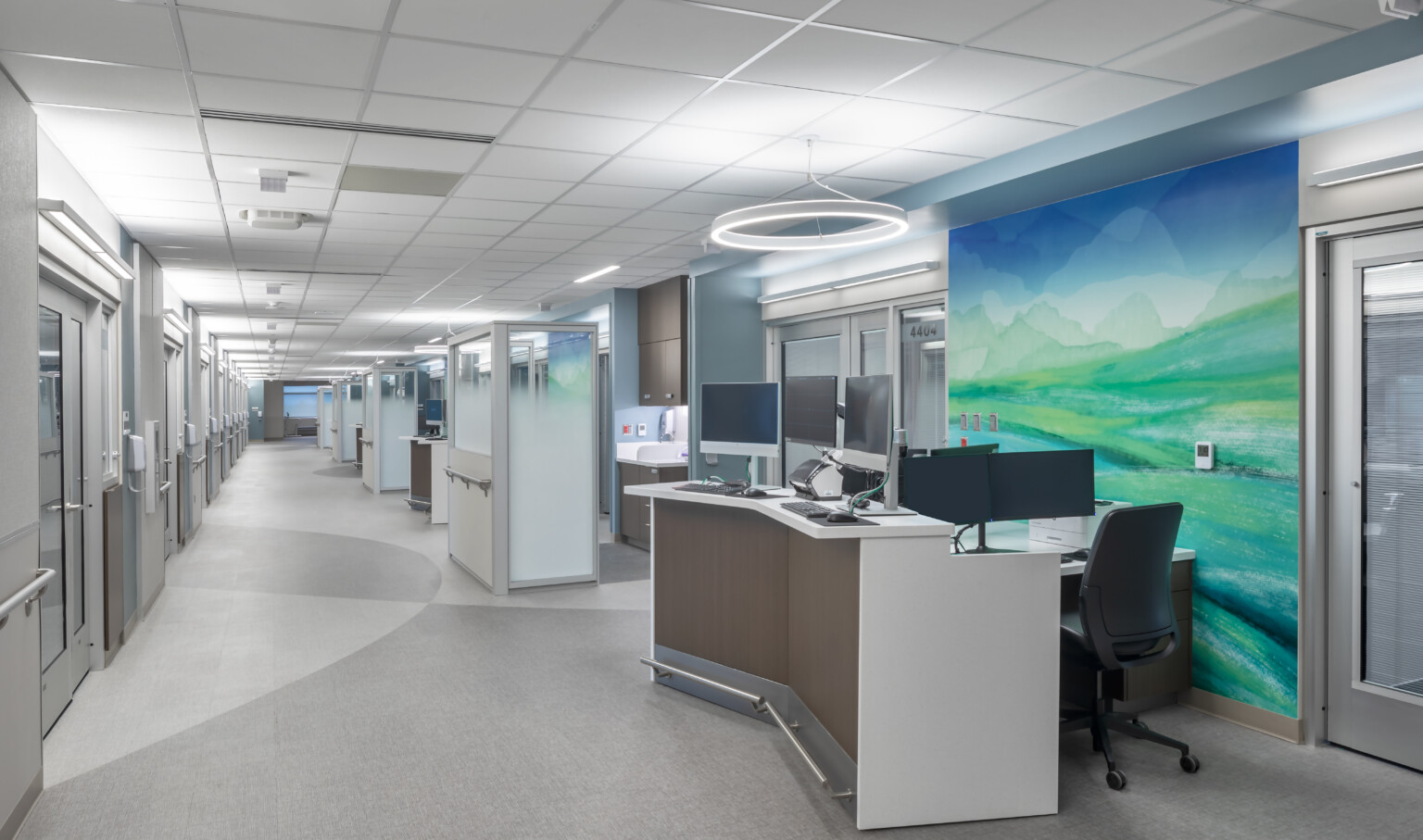 Hospital corridor with angled desk with multiple monitors in front of abstract blue and green mural. Glass partitions beyond Hospital corridor with angled desk with multiple monitors in front of abstract blue and green mural. Glass partitions beyond