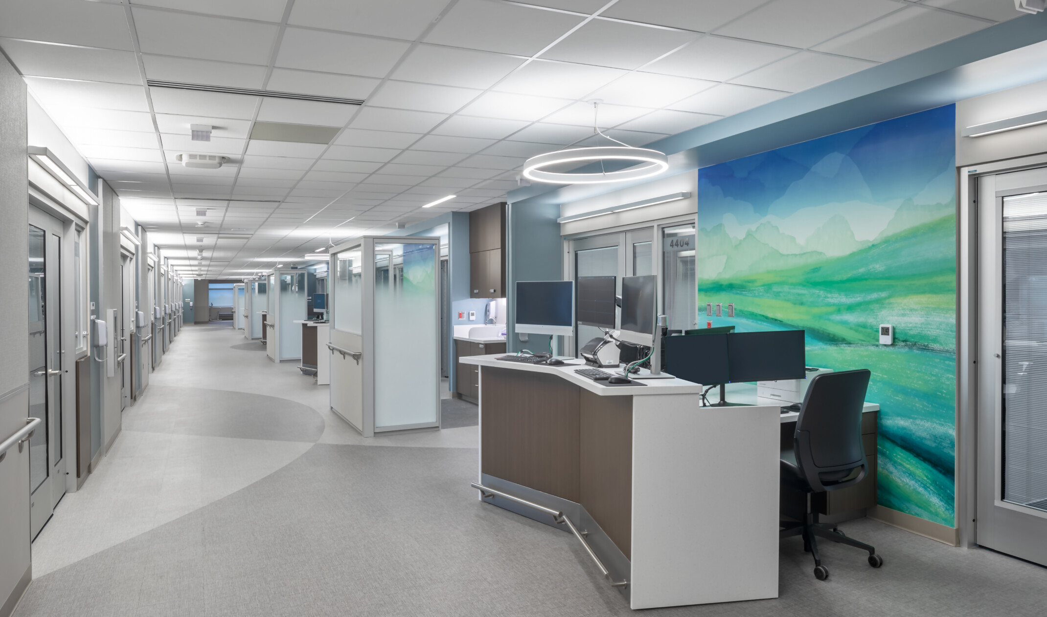 Hospital corridor with angled desk with multiple monitors in front of abstract blue and green mural. Glass partitions beyond