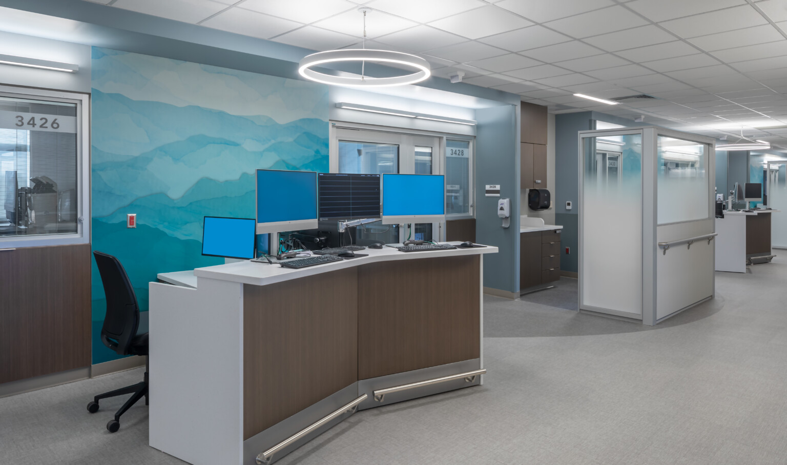Hospital corridor with angled desk with multiple monitors in front of abstract blue and green mural. Glass partitions beyond Hospital corridor with angled desk with multiple monitors in front of abstract blue and green mural. Glass partitions beyond