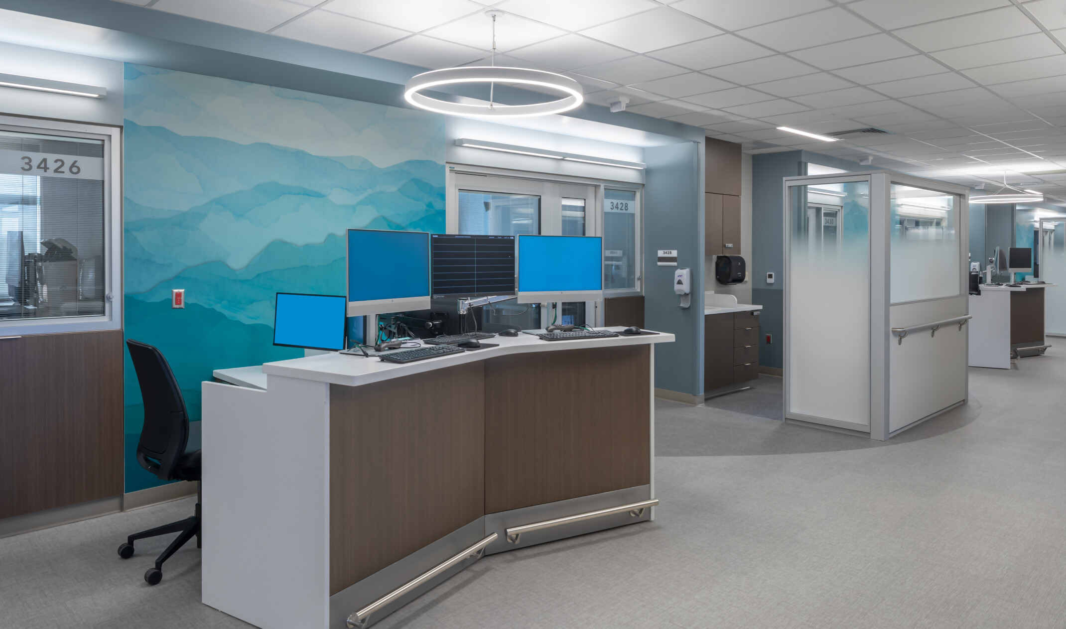 Hospital corridor with angled desk with multiple monitors in front of abstract blue and green mural. Glass partitions beyond