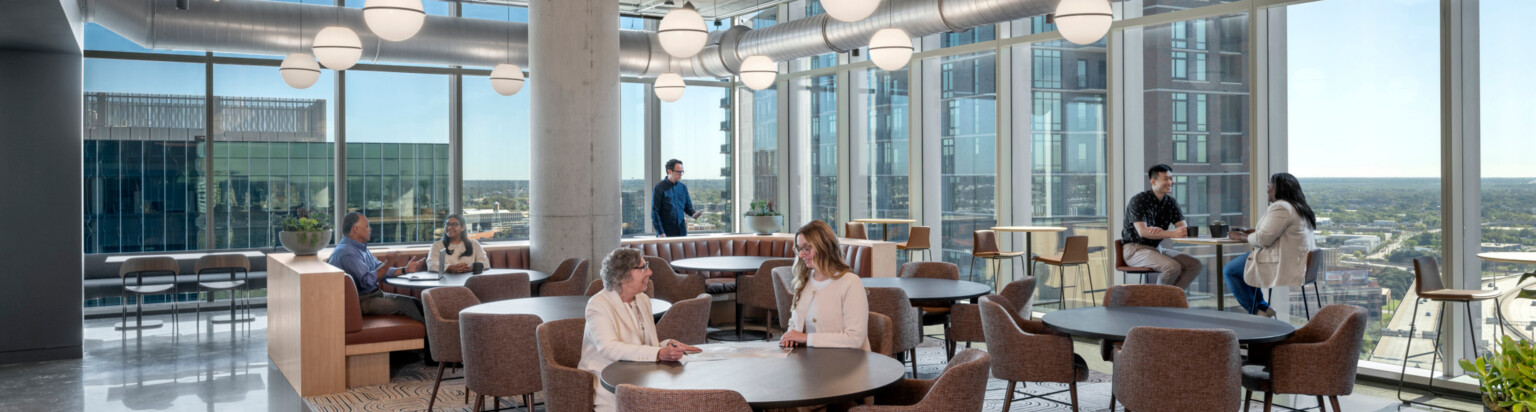 modern office collaboration hub with varied seating and people working at tables, booths and high tops in front of floor to ceiling windows; DLR Group Dallas office