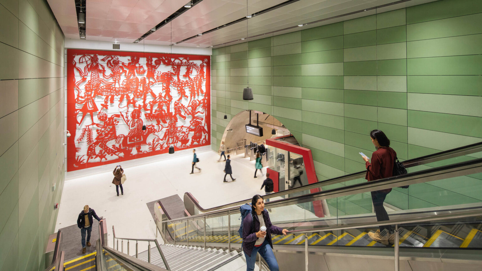 Concrete block walls with raised red sculptural mural on left. Right, arched hall with gates and ticket booth by escalators