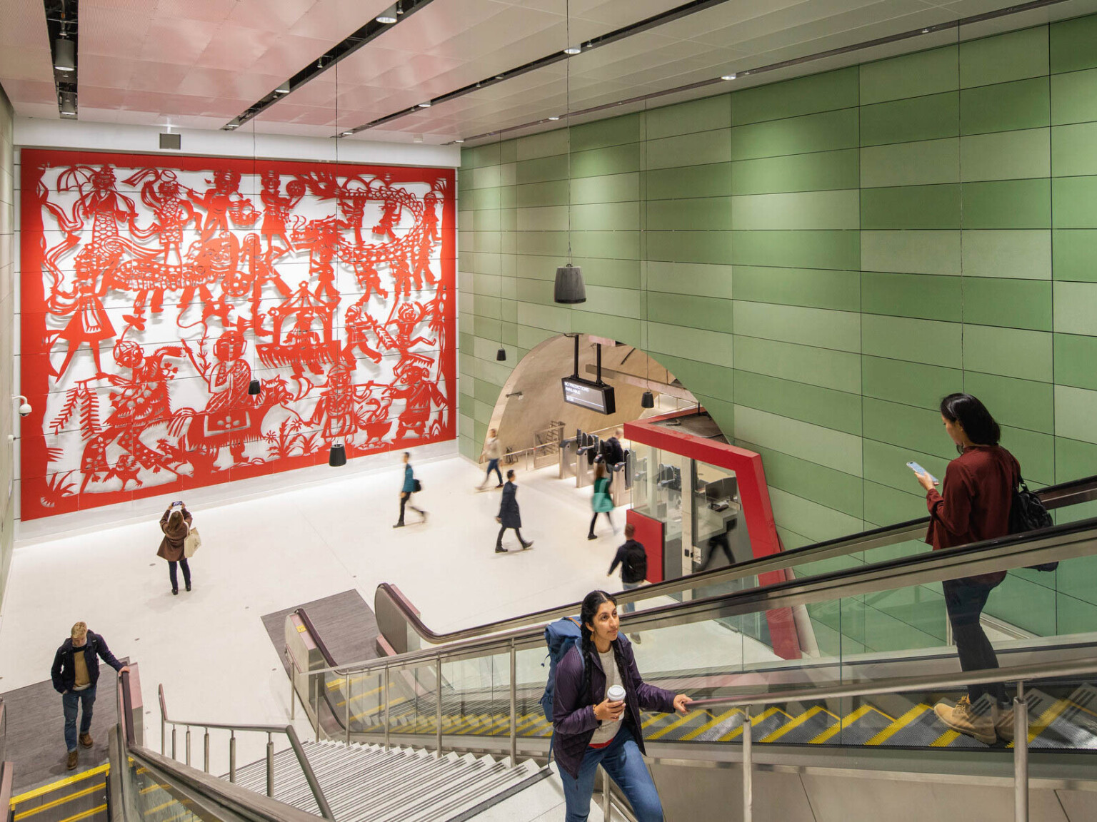 Concrete block walls with raised red sculptural mural on left. Right, arched hall with gates and ticket booth by escalators