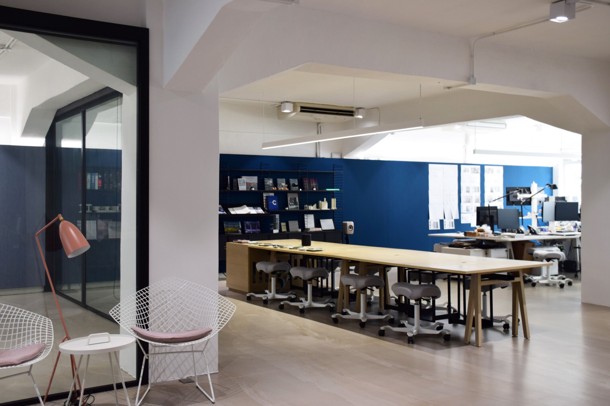 Interior of a modern office; large light wood table with stool seating; architecture and interior design workspaces in background