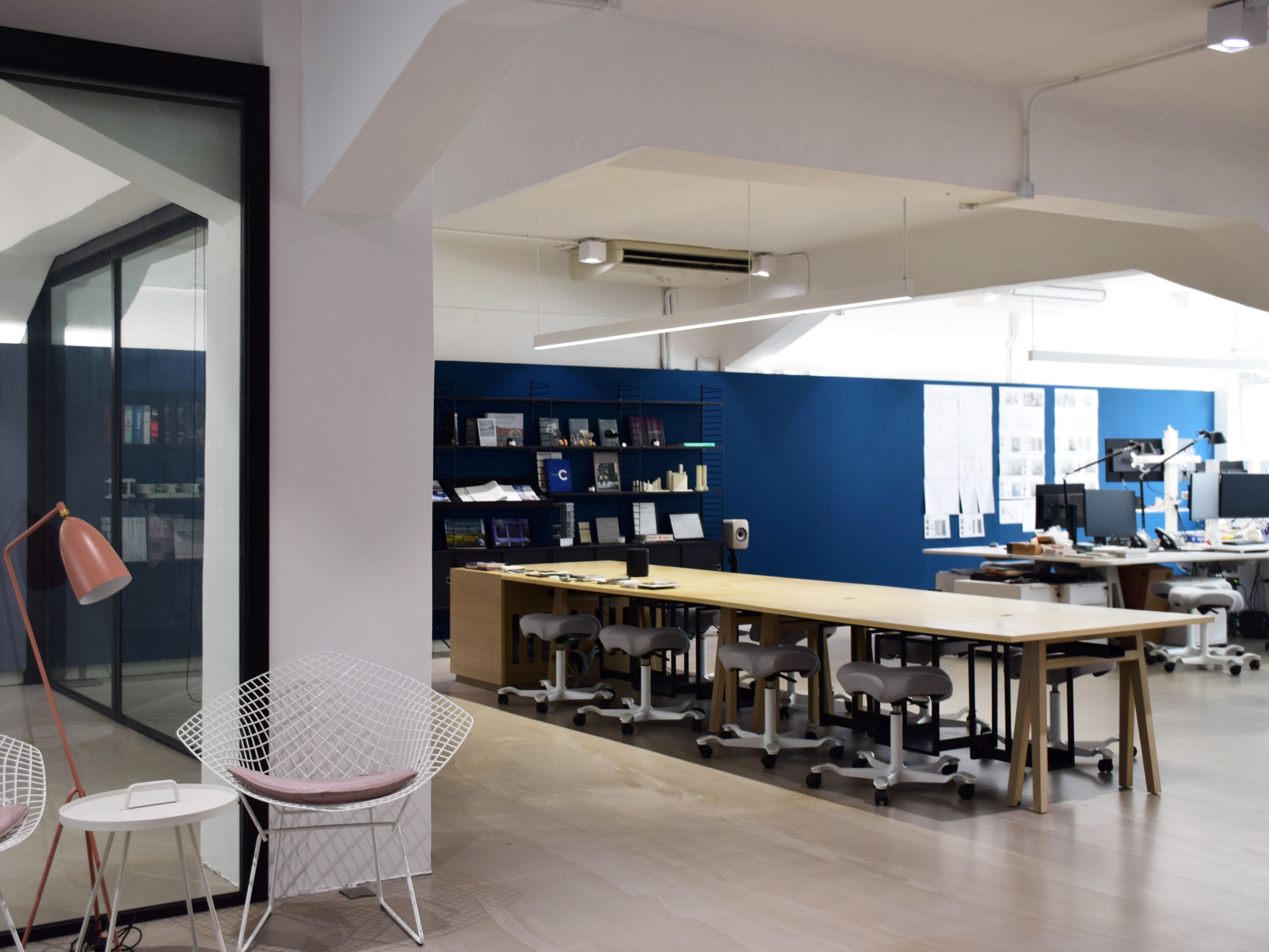 Interior of a modern office; large light wood table with stool seating; architecture and interior design workspaces in background
